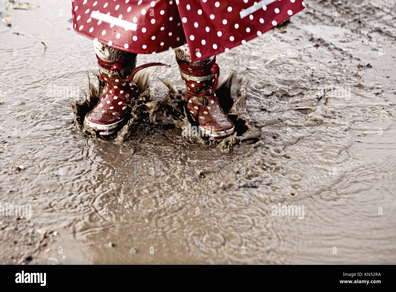 Detail of rainboots of a baby girl dressed with dotted raincoat in the puddles. Stock Photo