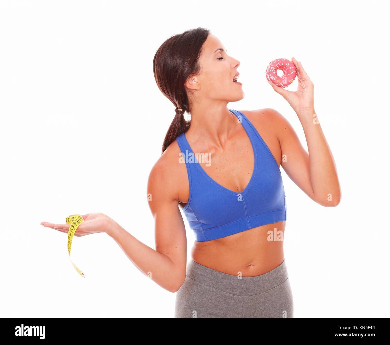 Fit woman in sport clothing holding measuring tape while biting a cake against white background. Stock Photo