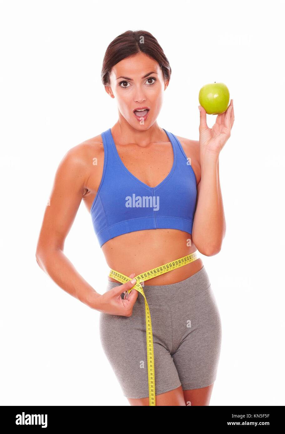 Slim female in sportswear taking waist measurement with measuring tape while looking at you on isolated background. Stock Photo