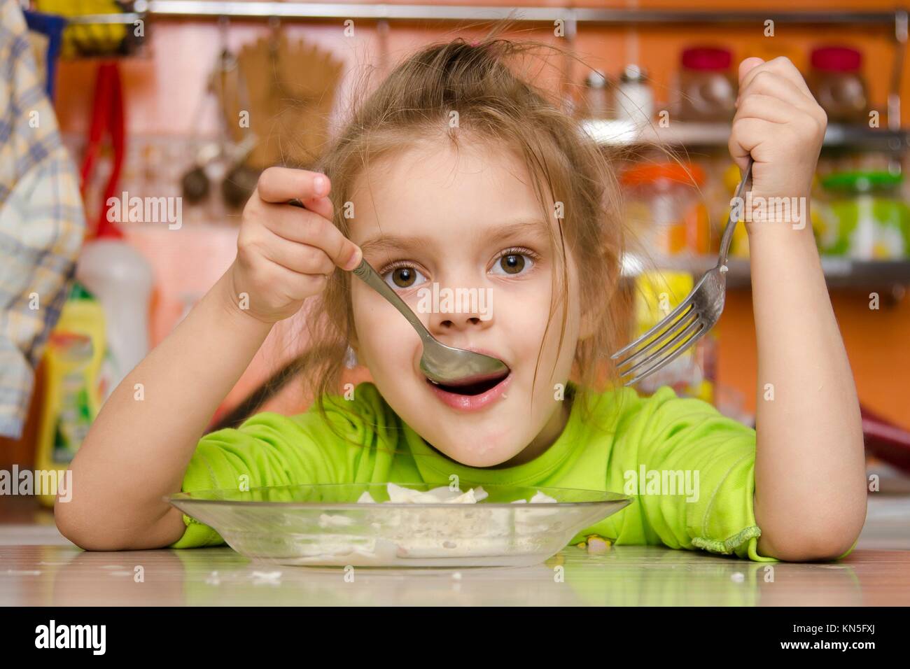A four year old girl eats sitting at the table in the kitchen. In one