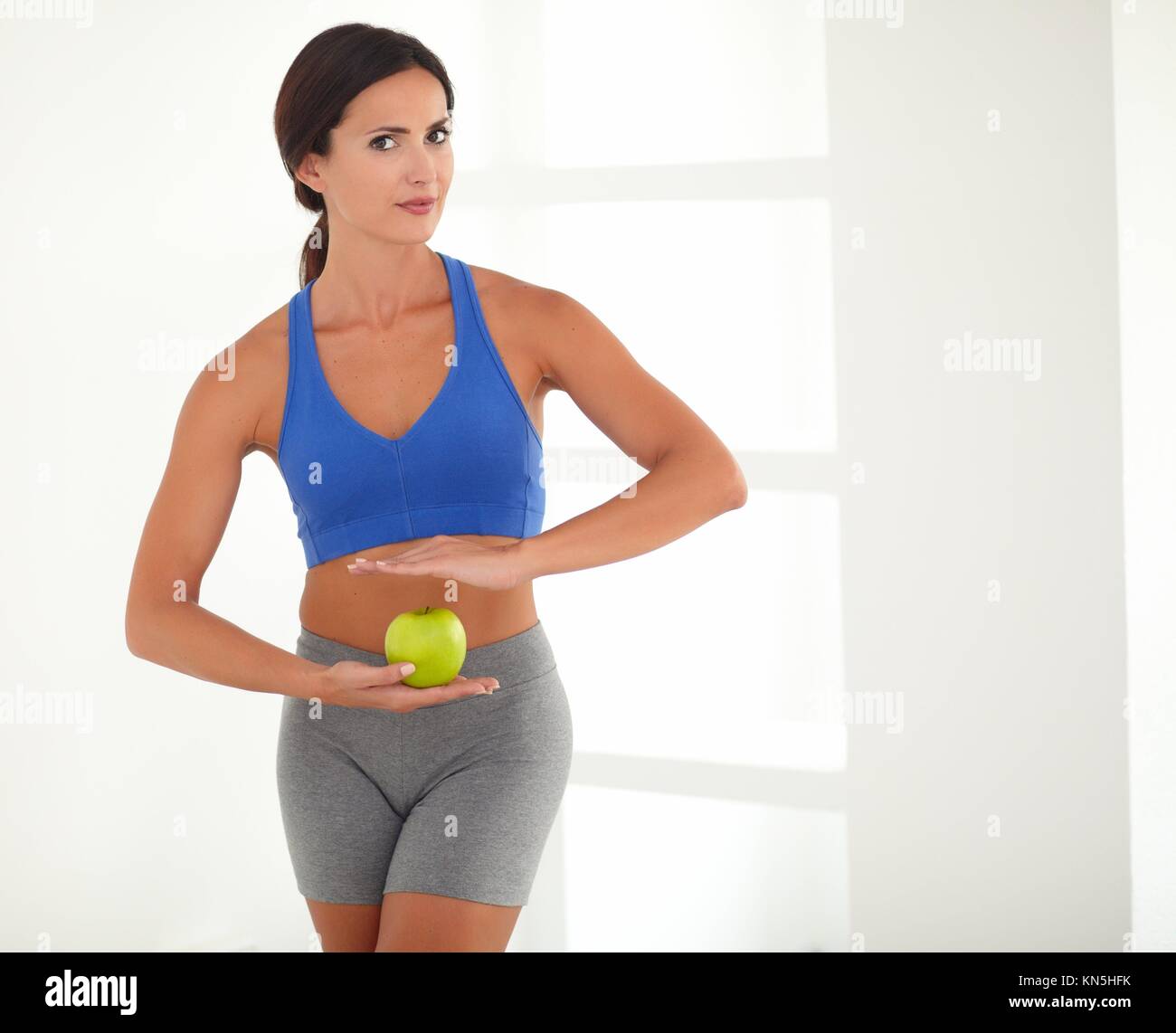 Hispanic woman in sports clothing holding apple while looking at you. Stock Photo