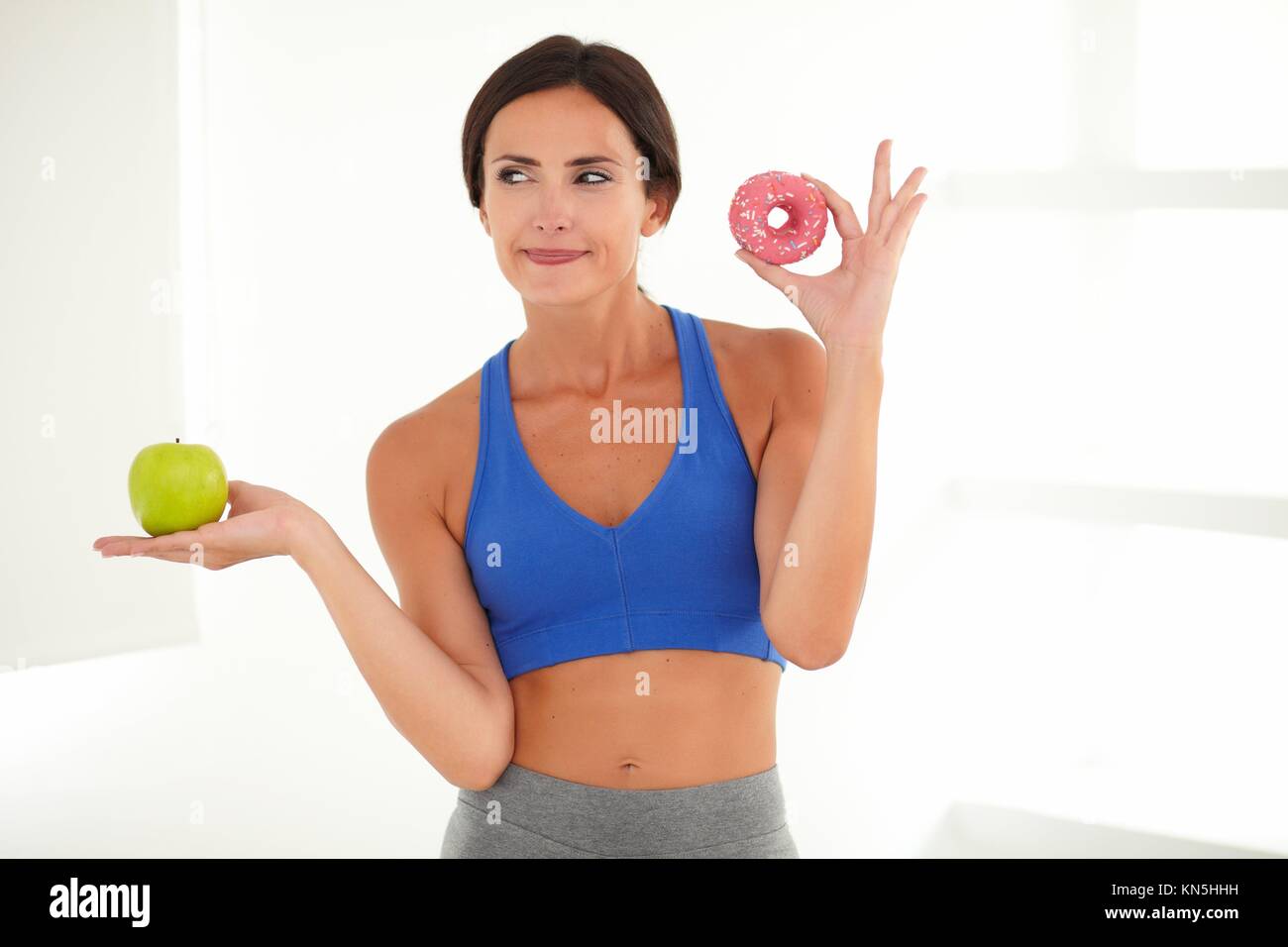 Female in training clothes holding sugary cake and fruit while is deciding. Stock Photo