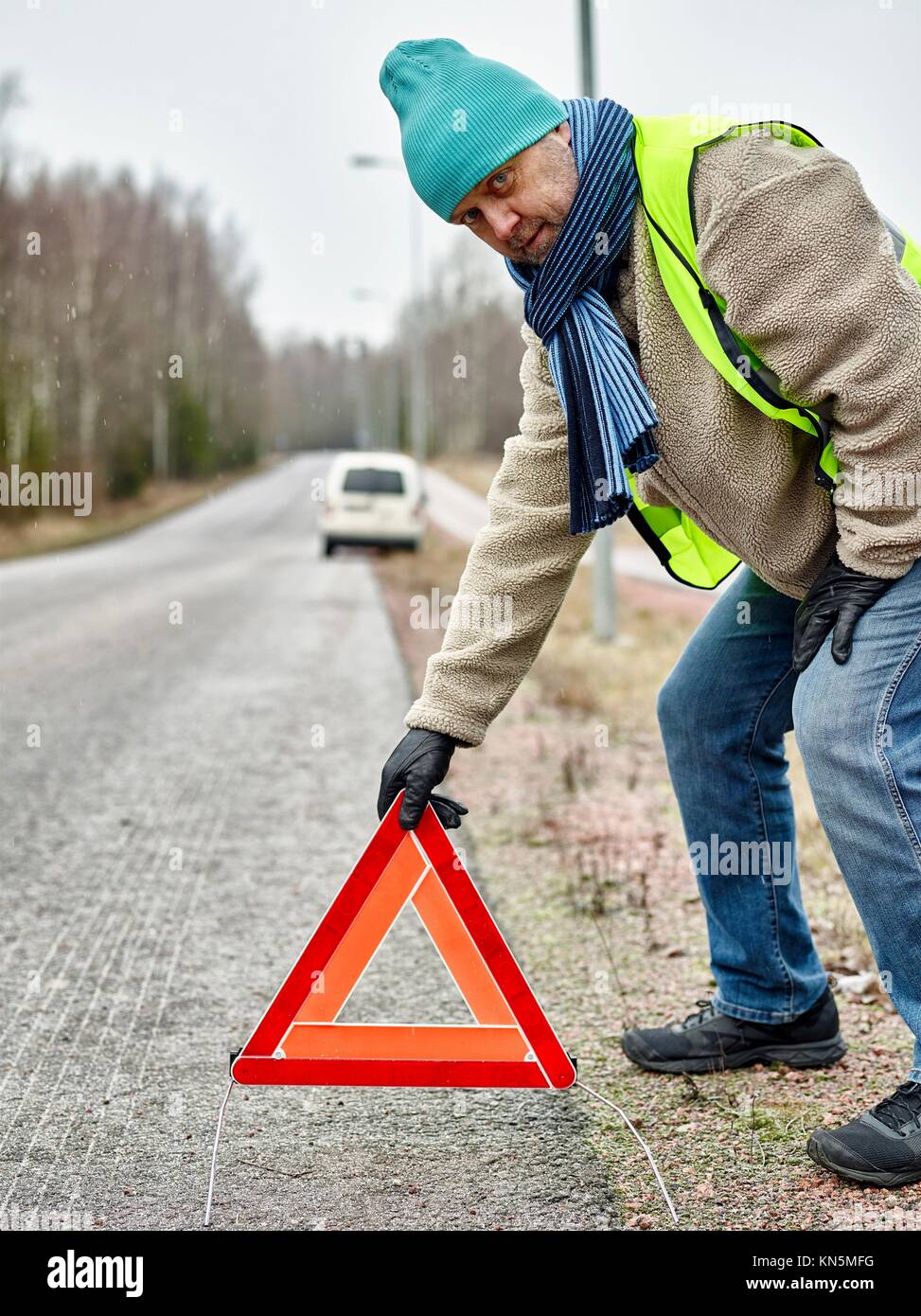 Mid adult man wearing reflector vest and he have a red warning triangle - road and vehicle on background. Stock Photo