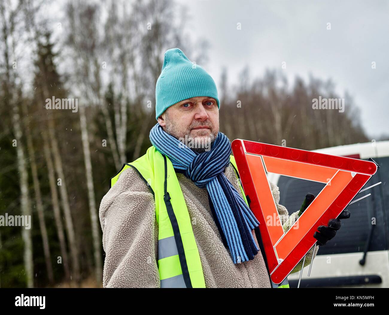 Mid adult man wearing reflector vest and he have a red warning triangle - vehicle on background. Stock Photo