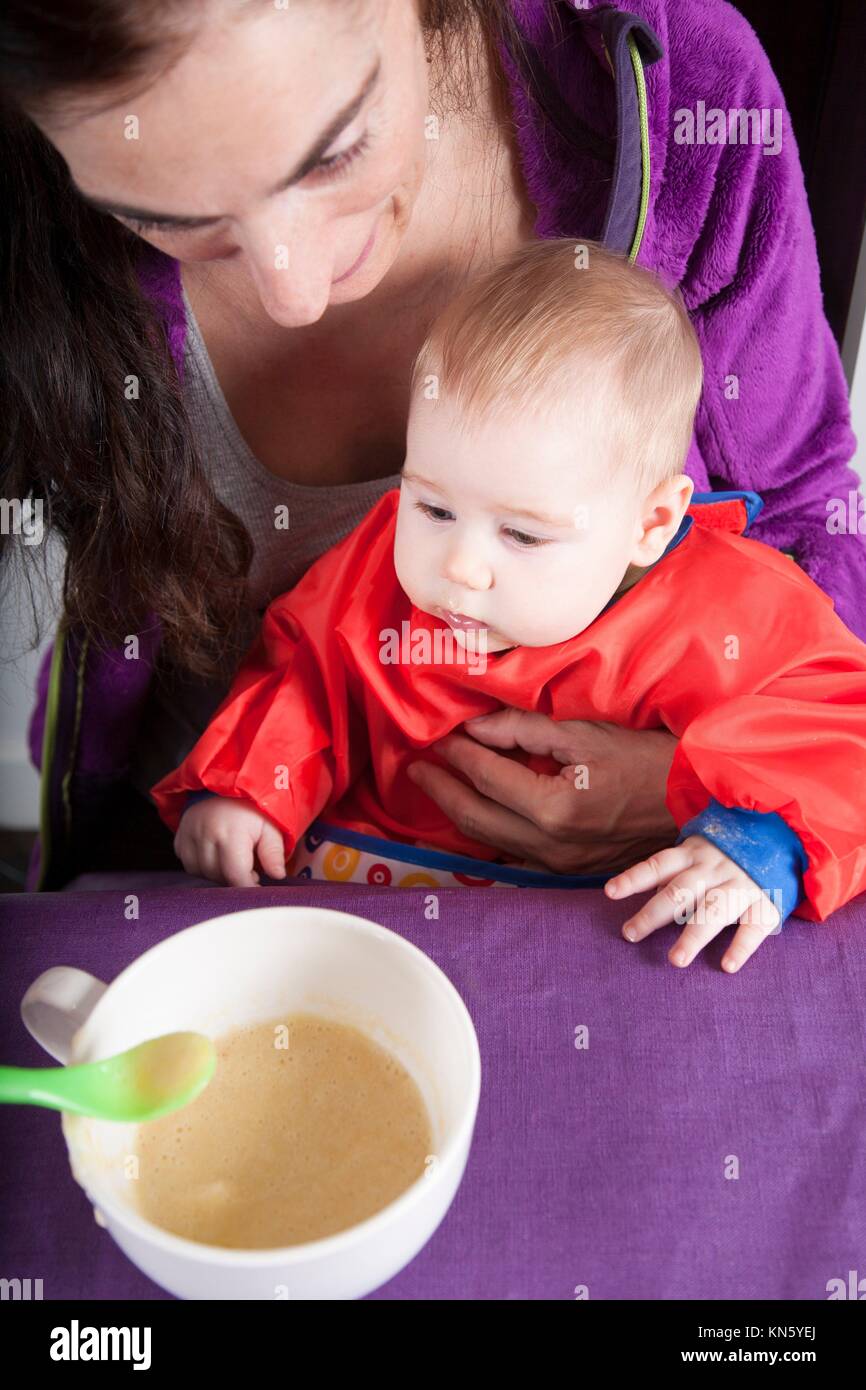 six months age blonde caucasian baby red bib in woman mother purple velvet jacket arms eating puree. Stock Photo