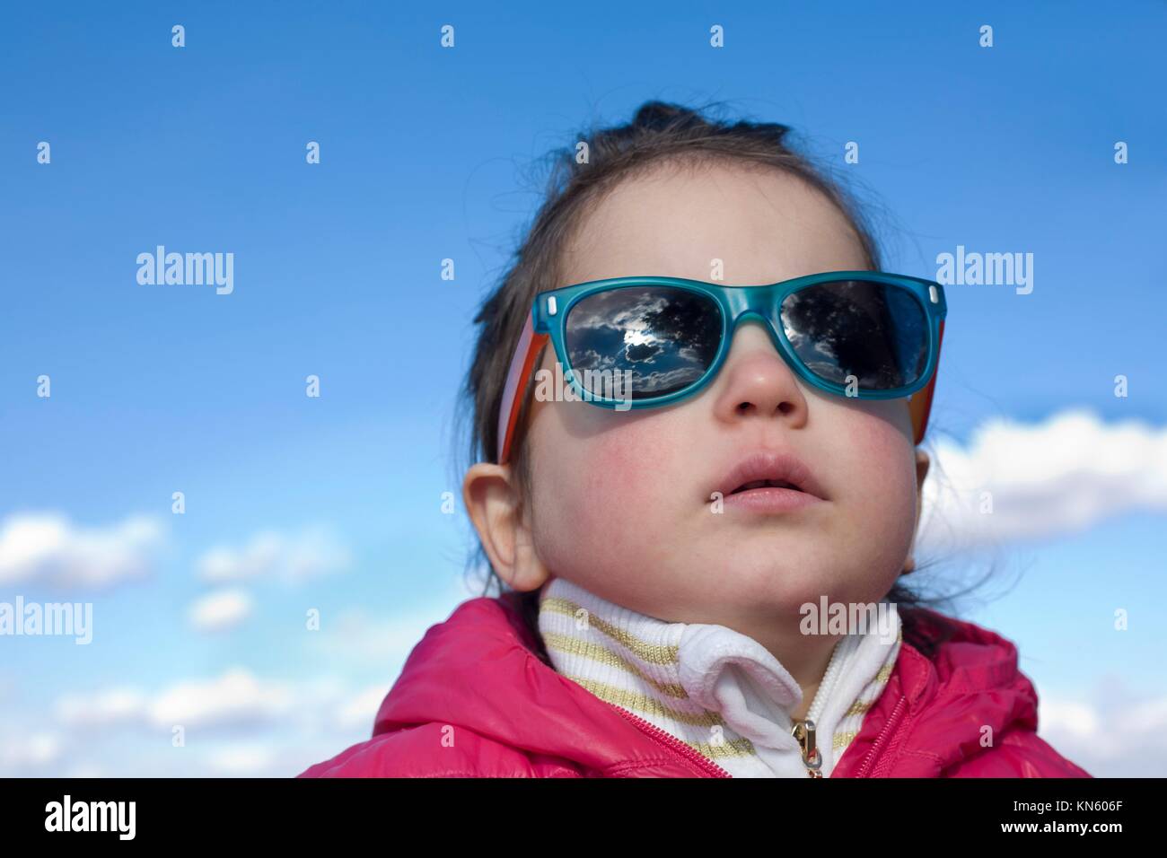 Portrait of cute 2,5 years old toddler girl with fashion children sunglasses with coat over blue sky with clouds background. Stock Photo
