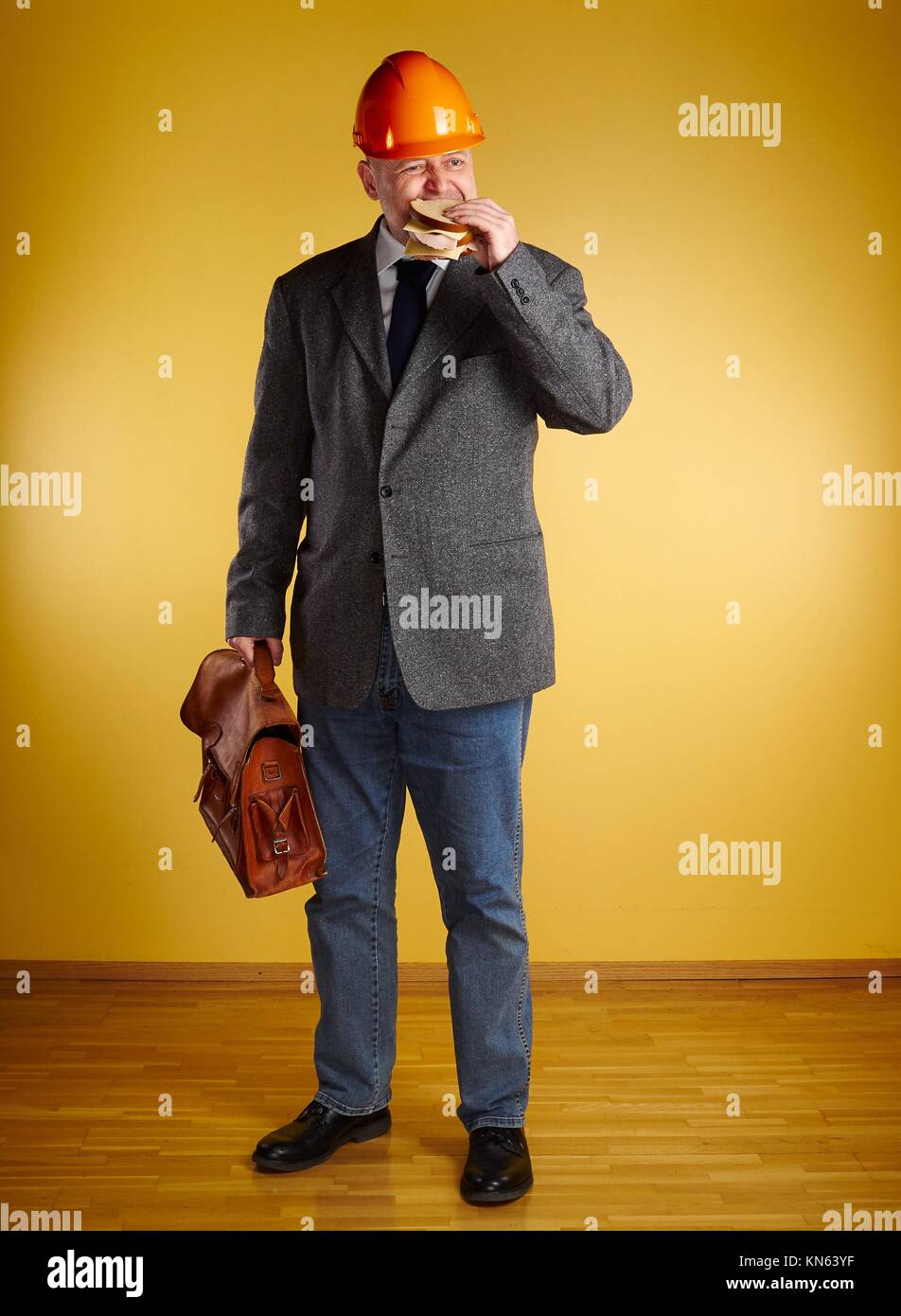 Male engineer in room, he eats sandwich and holds old leather briefcase, parquet floor and yellow wall. Stock Photo