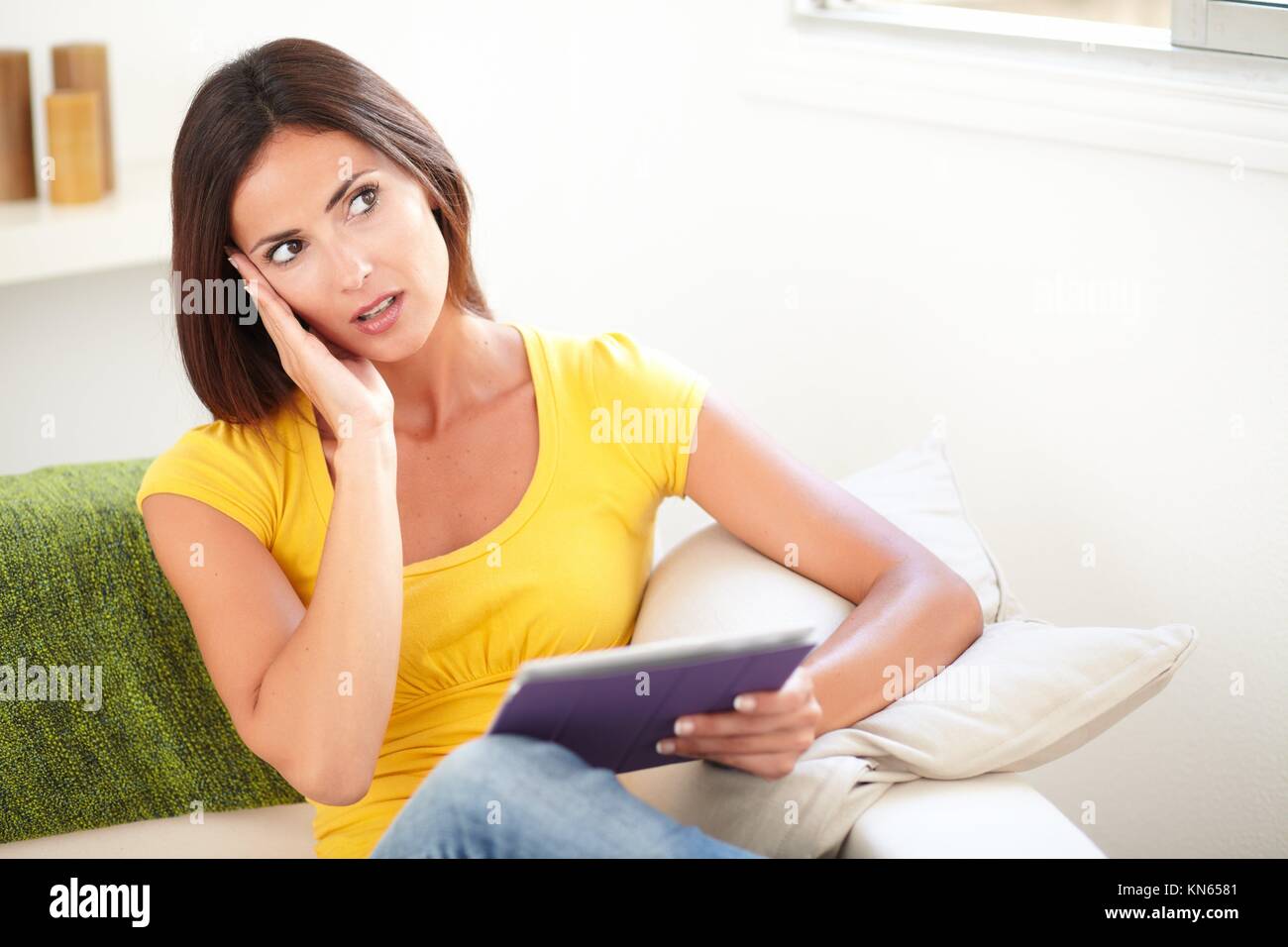 Surprised lady in yellow tank top holding a tablet while looking away from the camera - copy space. Stock Photo