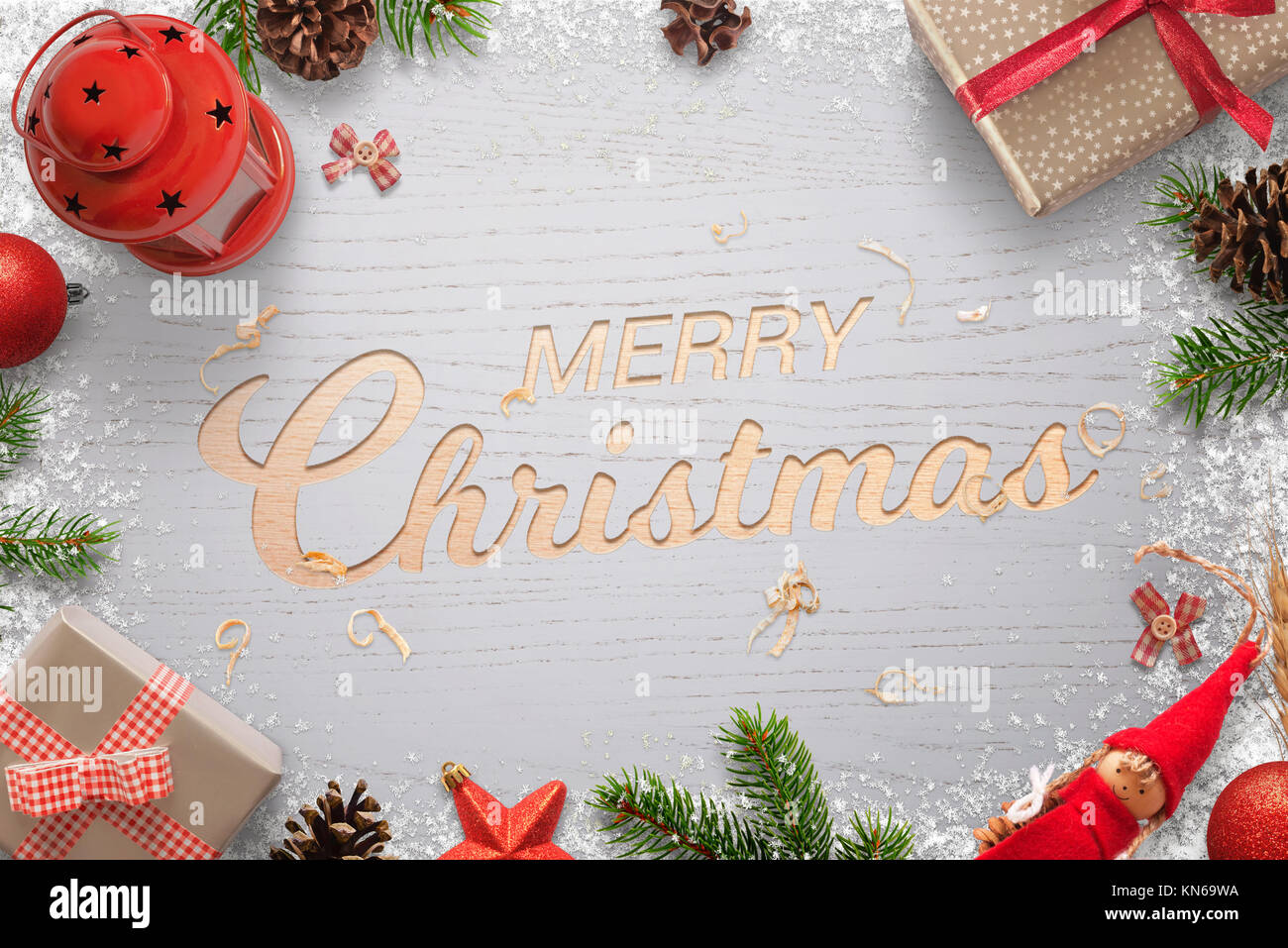 Merry Christmas text carved in a wooden surface and surrounded by Christmas decorations. Christmas tree, gifts, balls, lantern, pinecones and doll. Stock Photo