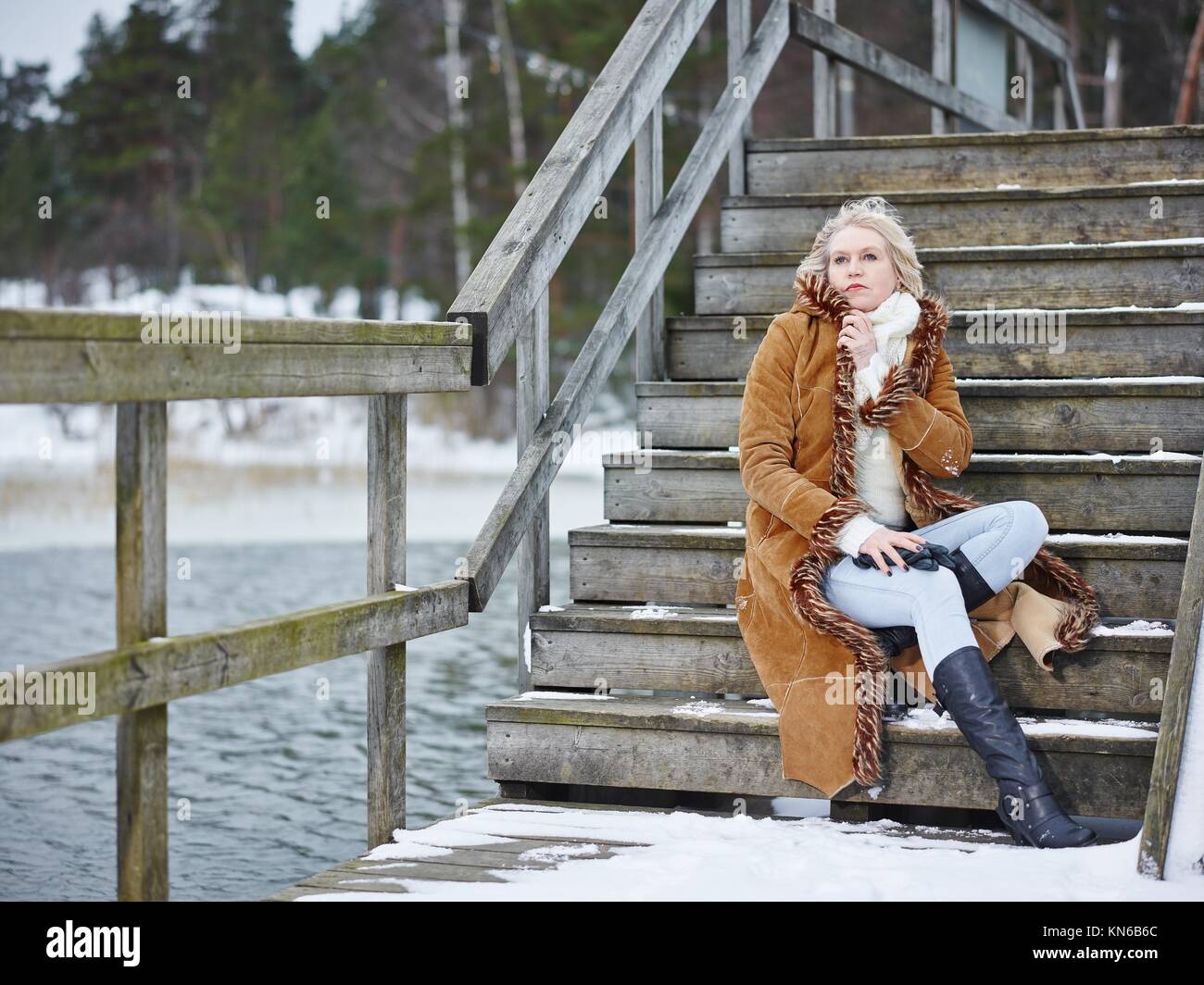 Fashionable mature adult woman wearing winter clothes and she sitting on the stairs - rural scene. South Finland in January. Stock Photo