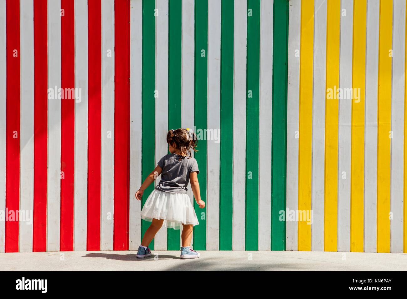 Three years old girl playing beside striped colored wall of several old beach huts. Stock Photo