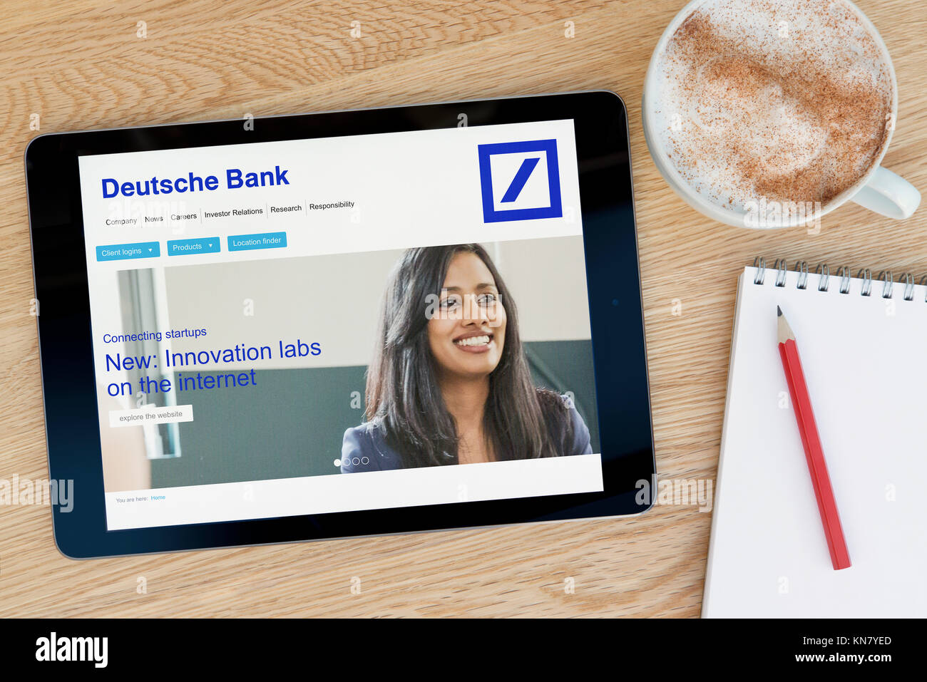 The Deutsche Bank website on an iPad tablet device which rests on a wooden table beside a notepad and pencil and a cup of coffee (Editorial only) Stock Photo