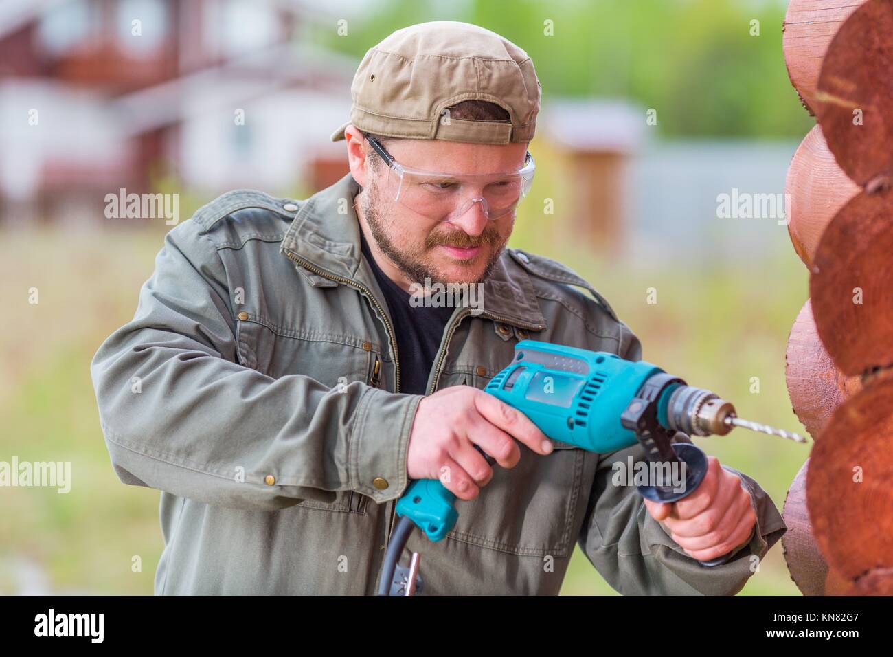 Worker with a drill in safety glasses working in a wooden cottage. Stock Photo