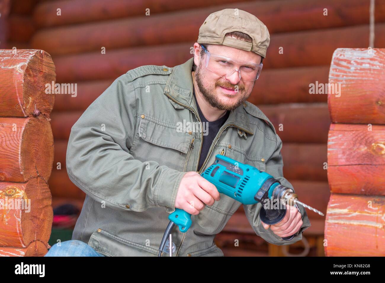 Worker with a drill in safety glasses working in a wooden cottage. Stock Photo