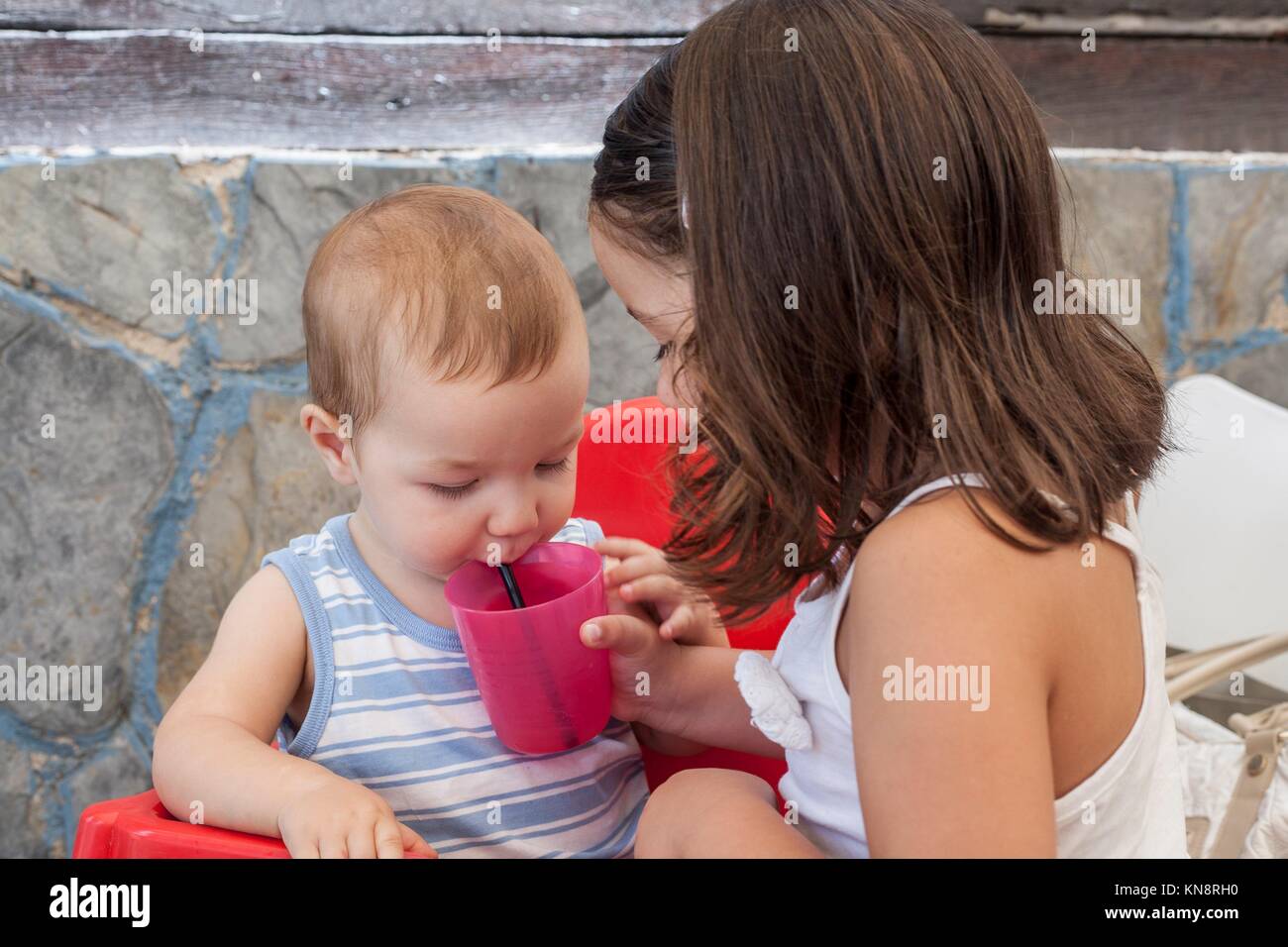 Little sister helping to her baby brother to drink juice with straw. She is holding the plastic glass. Stock Photo