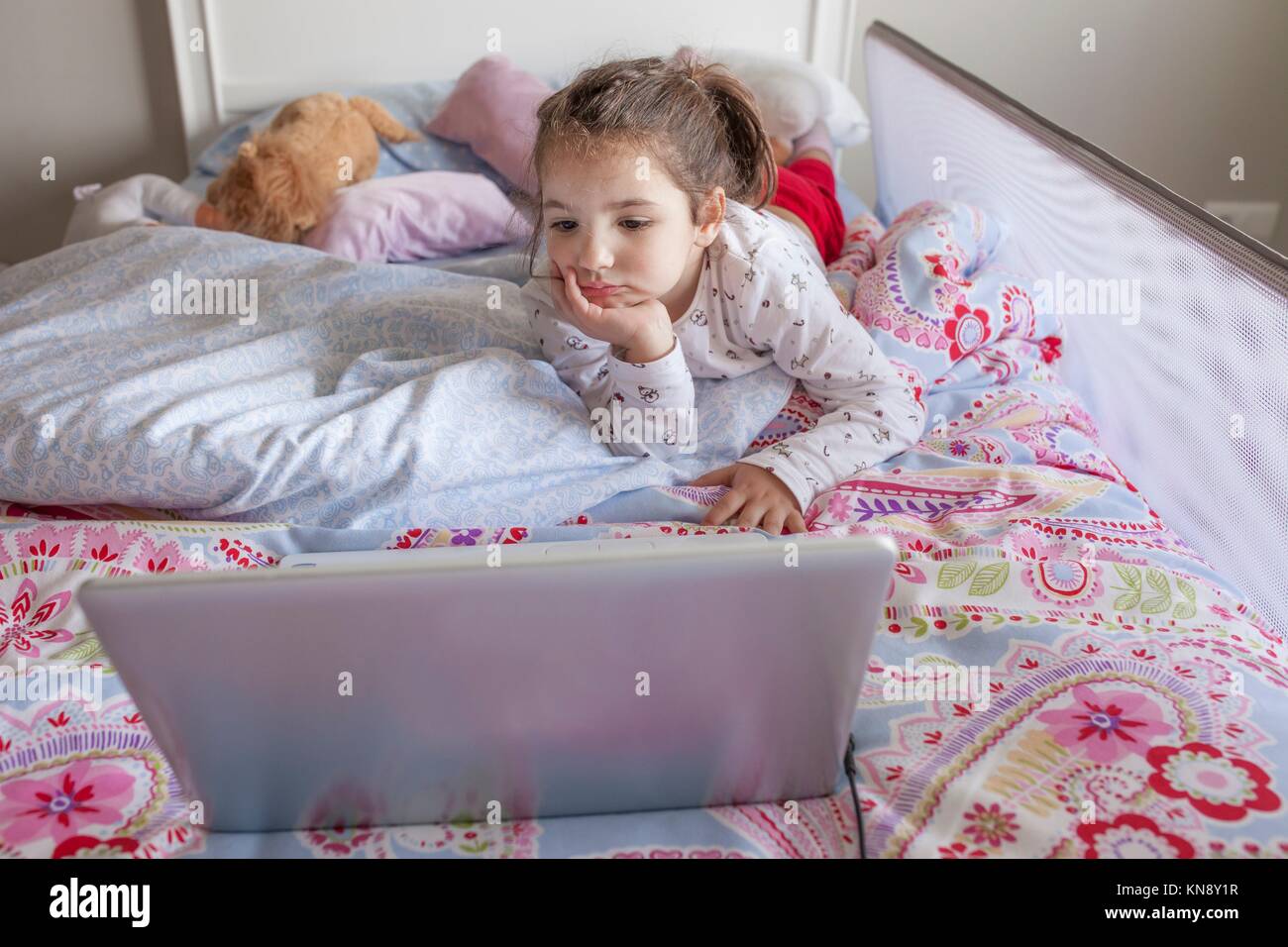Little girl lying in bed and watching cartoons with a laptop in his bedroom. She looks entertained. Stock Photo