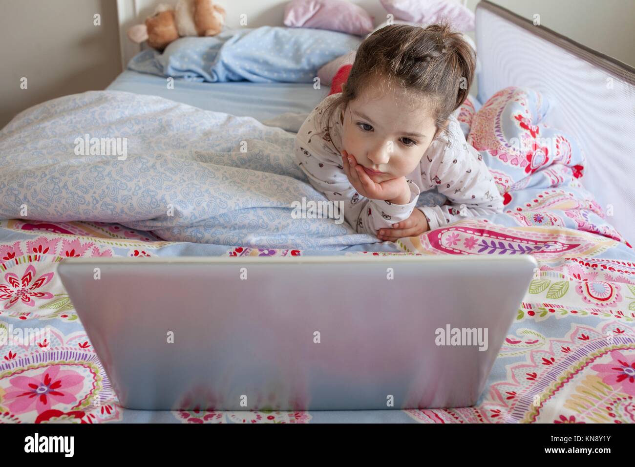 Little girl lying in bed and playing with a laptop in his bedroom. She looks attentive. Stock Photo