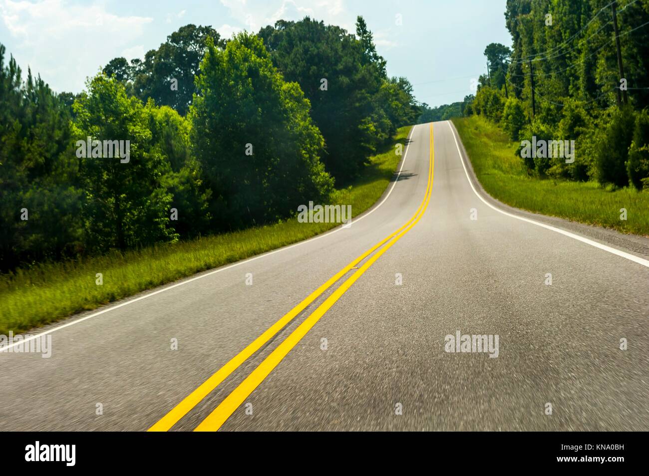 A 2 lane paved country road with double yellow lines and trees on both