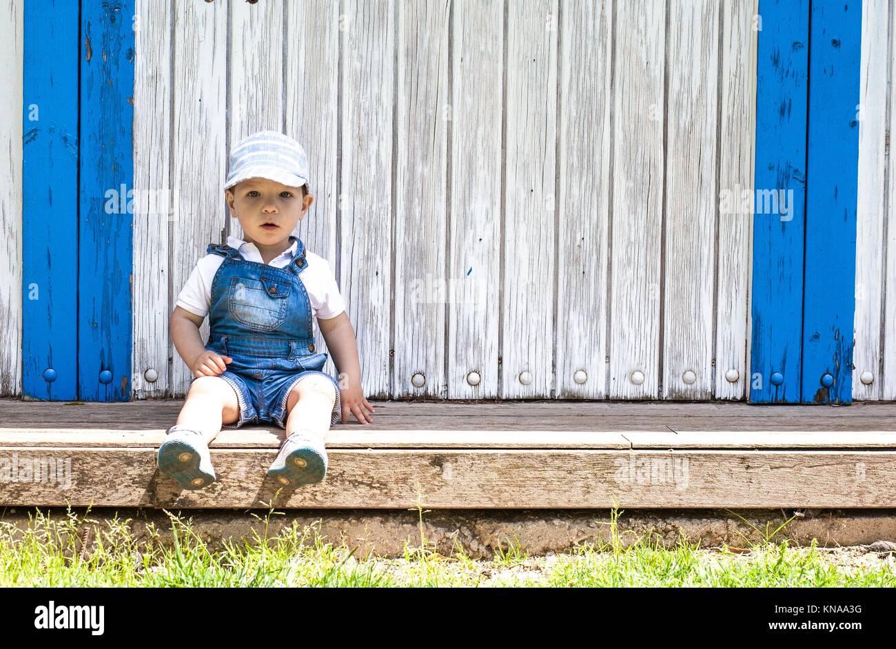2 year-old boy sitting portrait at wooden beach hut. He wears a cap. Stock Photo