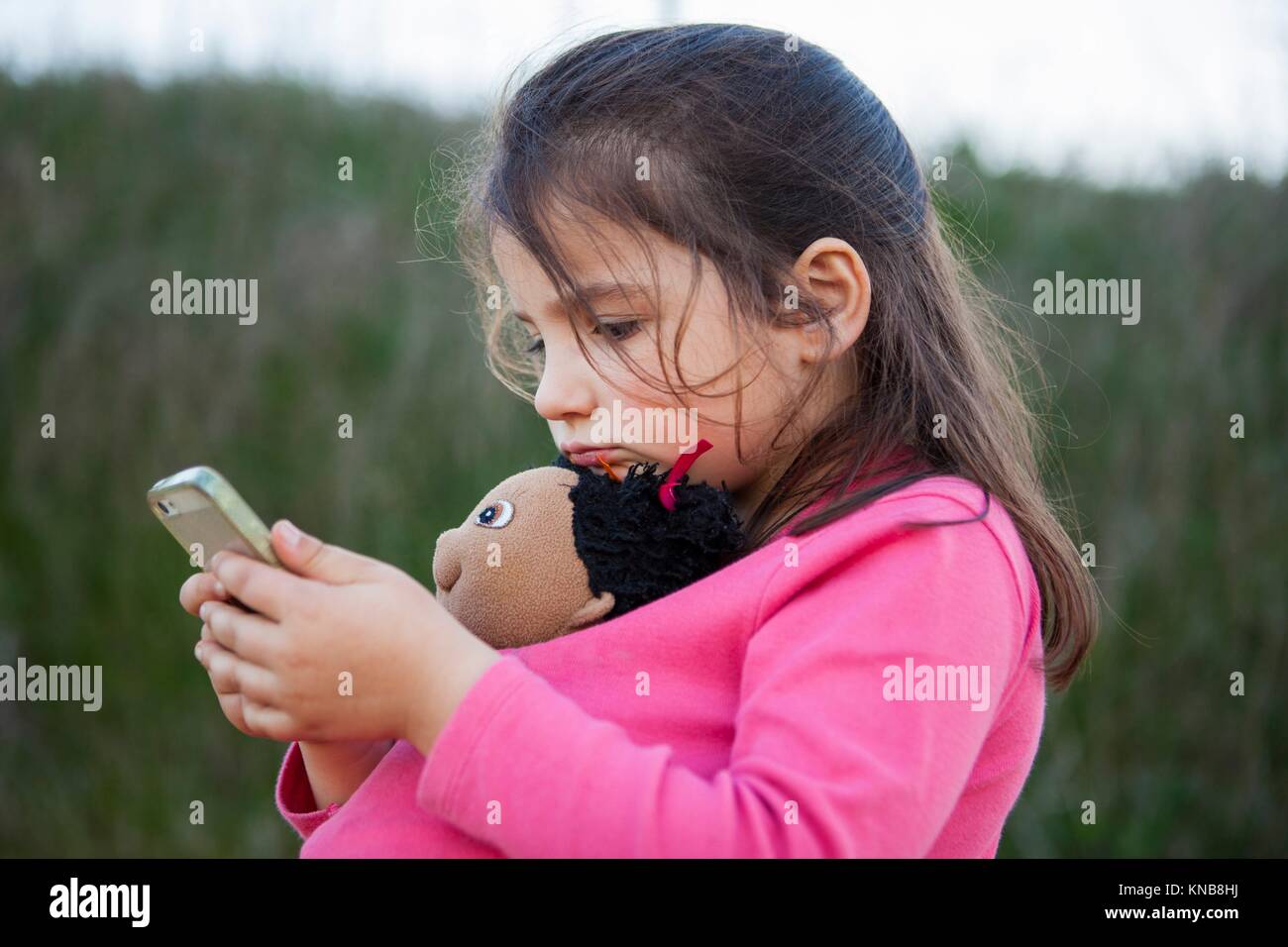 Little girl playing with mobile phone and holding her doll outdoor. Stock Photo