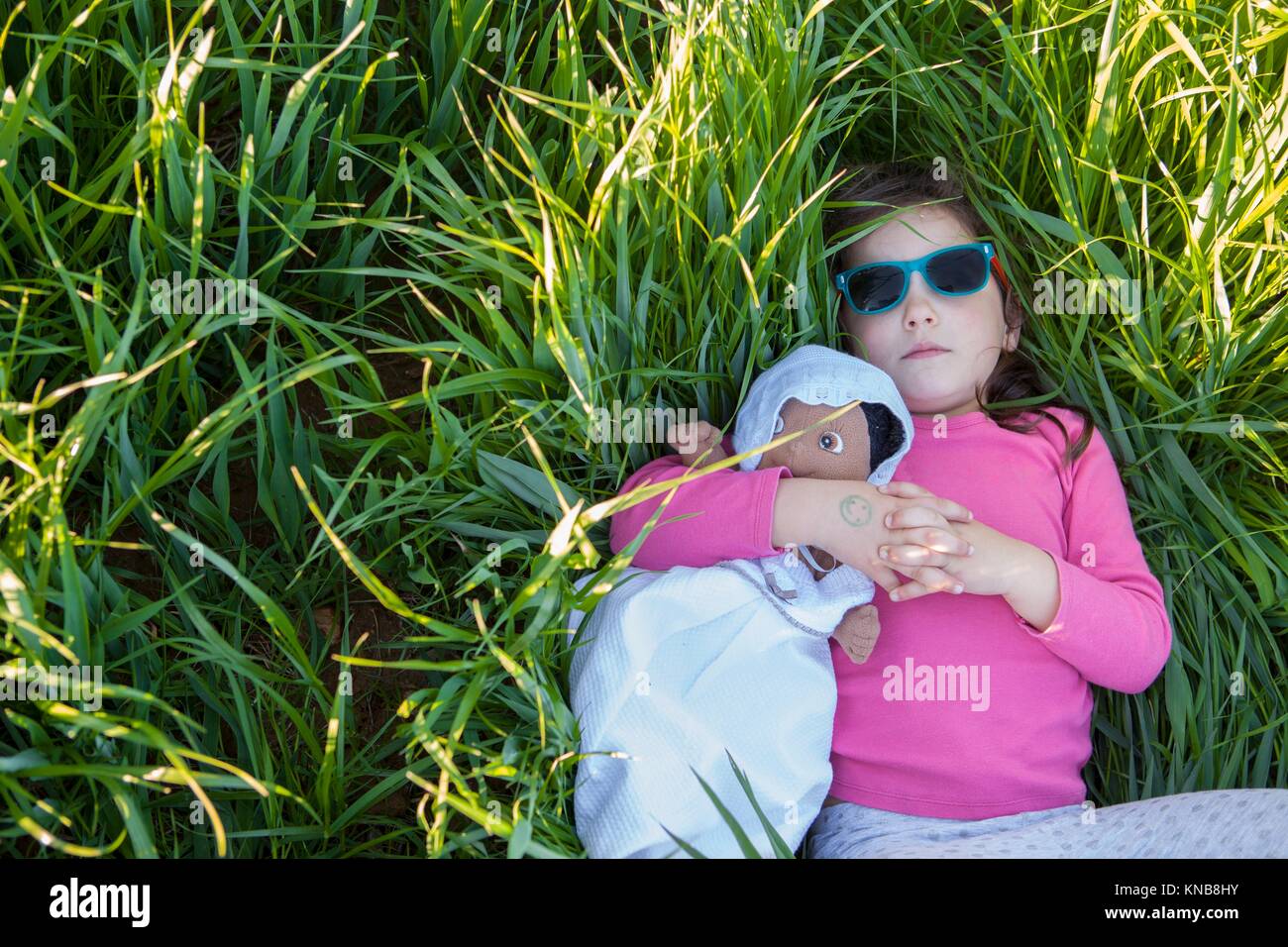 Little girl and her doll lying on green cereal field at sunset, Spain. Stock Photo