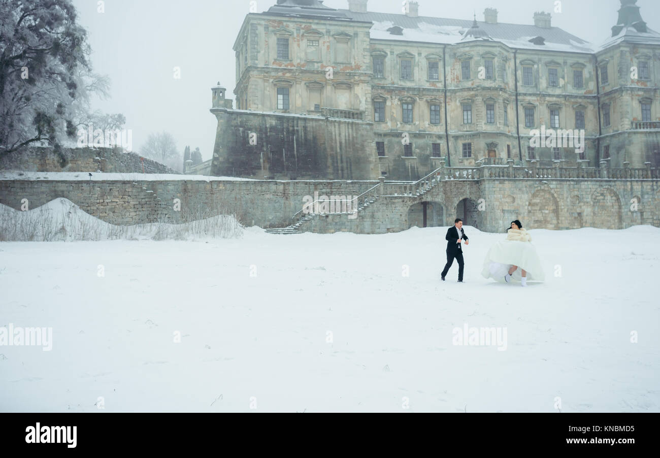 Newlyweds couple is having fun during their walk on the snowy meadow near the old house. The groom is joyfully running after the beautiful bride. Stock Photo