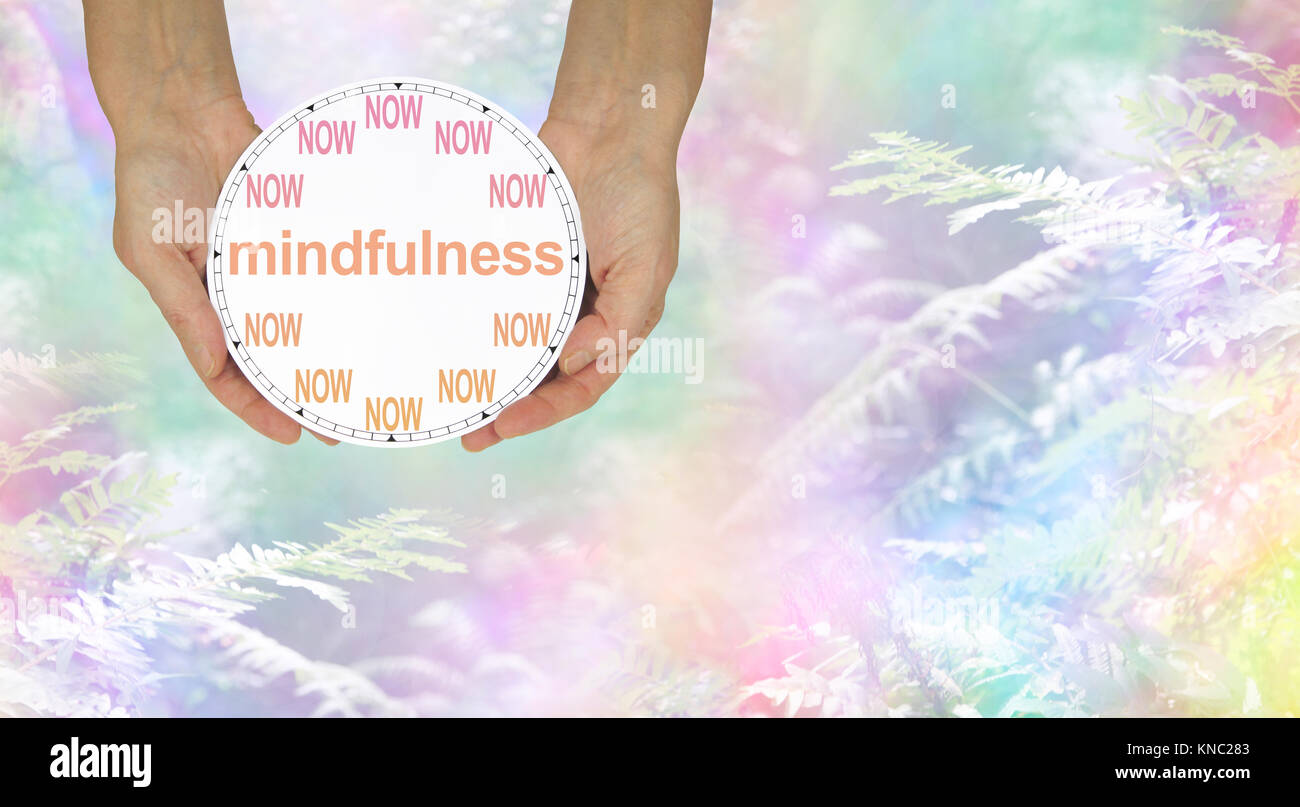 Mindfulness - do it NOW - female hands holding a NOW clock with MINDFULNESS written across the middle on rainbow coloured nature background Stock Photo