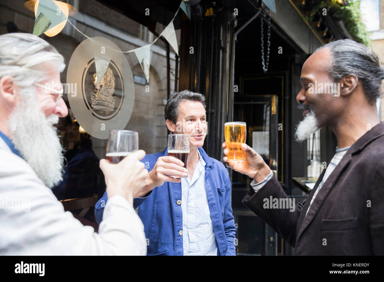 Three mature men, standing outside pub, holding beer glasses, making a toast Stock Photo