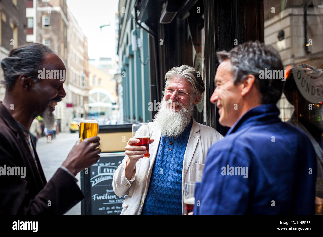 Three mature men, standing outside pub, holding beer glasses, laughing Stock Photo