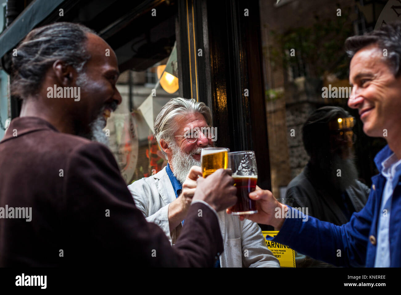 Three mature men, standing outside pub, holding beer glasses, making a toast Stock Photo
