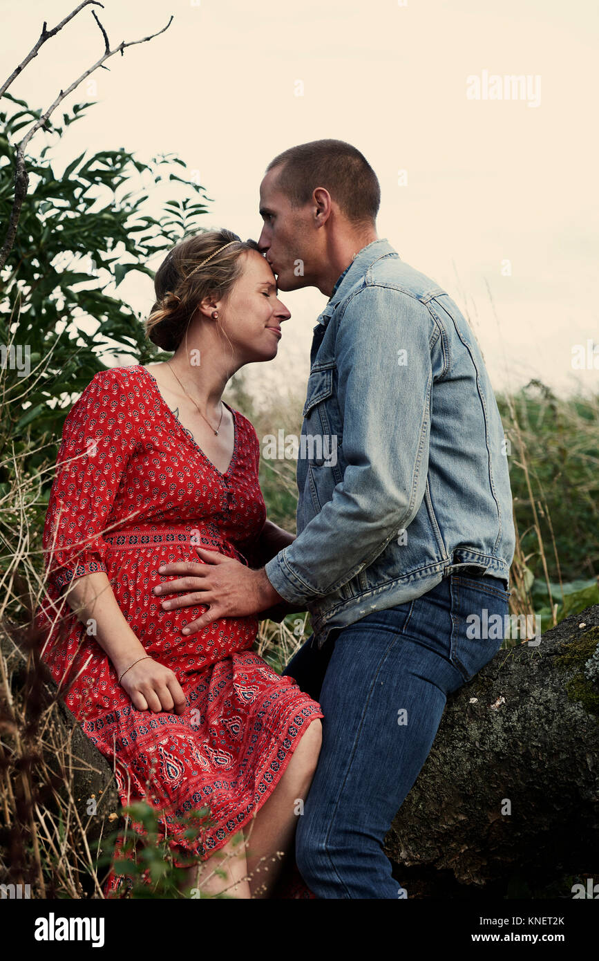 Romantic man kissing pregnant wife's forehead on tree trunk Stock Photo