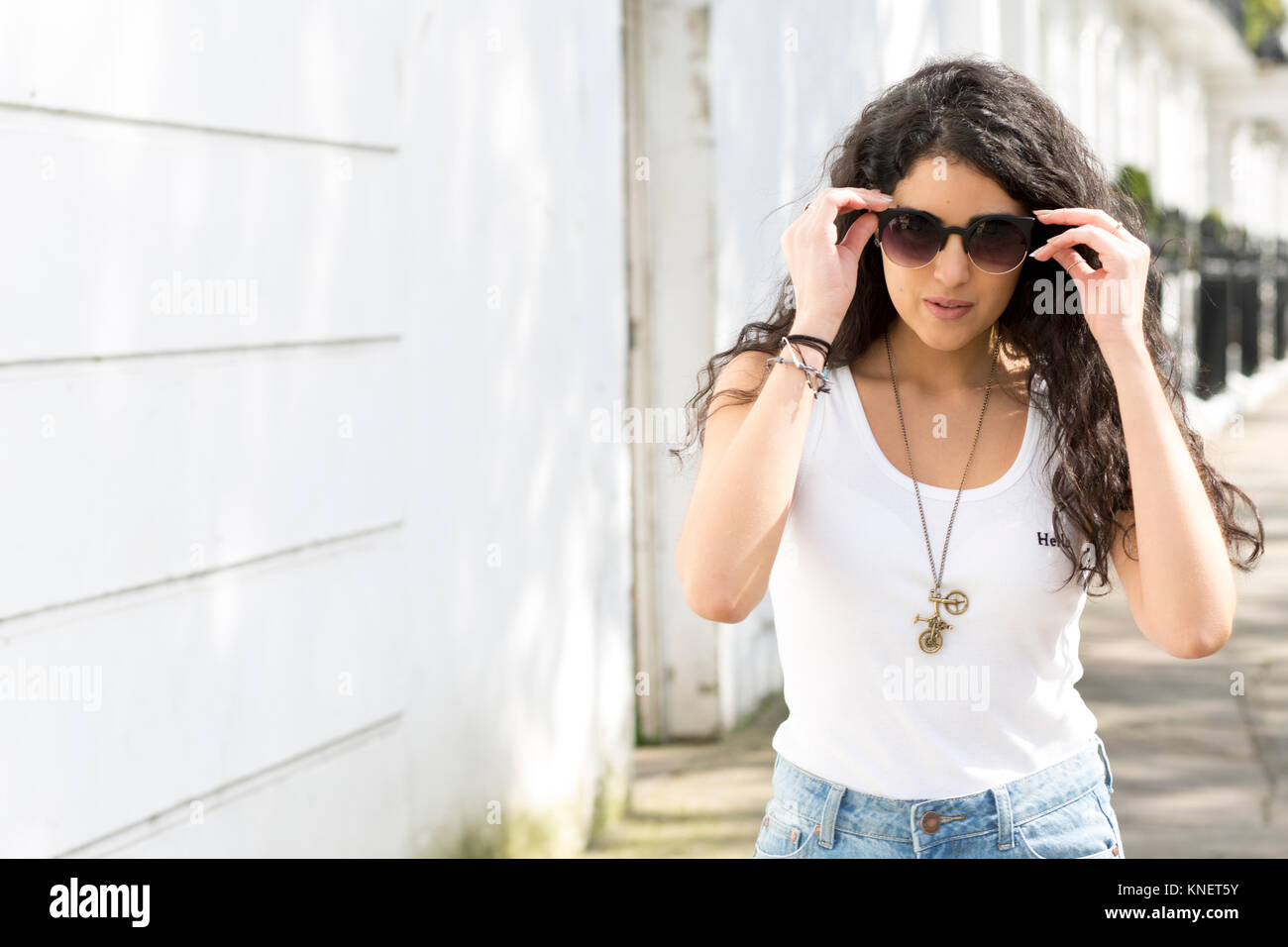 Young woman with long wavy hair strolling on street putting on sunglasses Stock Photo