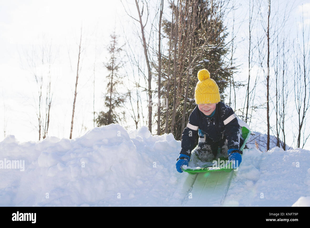 Boy in yellow knit hat tobogganing on snow covered hill Stock Photo