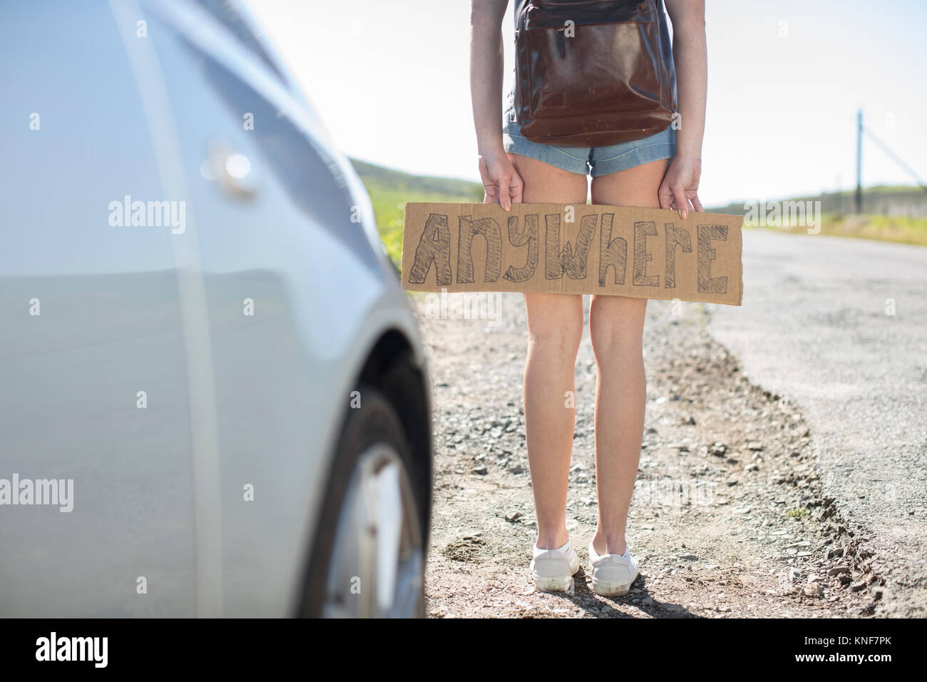 Young woman standing beside car, holding hitch-hiking sign saying 'anywhere', low section Stock Photo