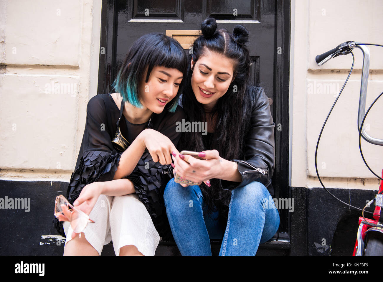 Two young stylish female friends sitting doorstep looking at smartphone Stock Photo
