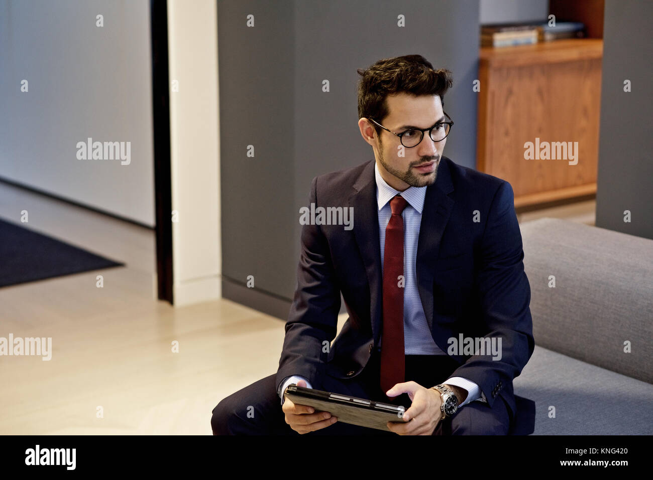 Latino businessman dressed in a smart suit holding a mobile tablet device in a contemporary interior designed office Stock Photo