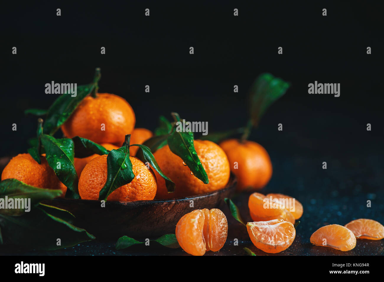 Citrus fruits on a wooden plate with green leaves. Vibrant tangerines on a dark background. Rustic food photography. Stock Photo