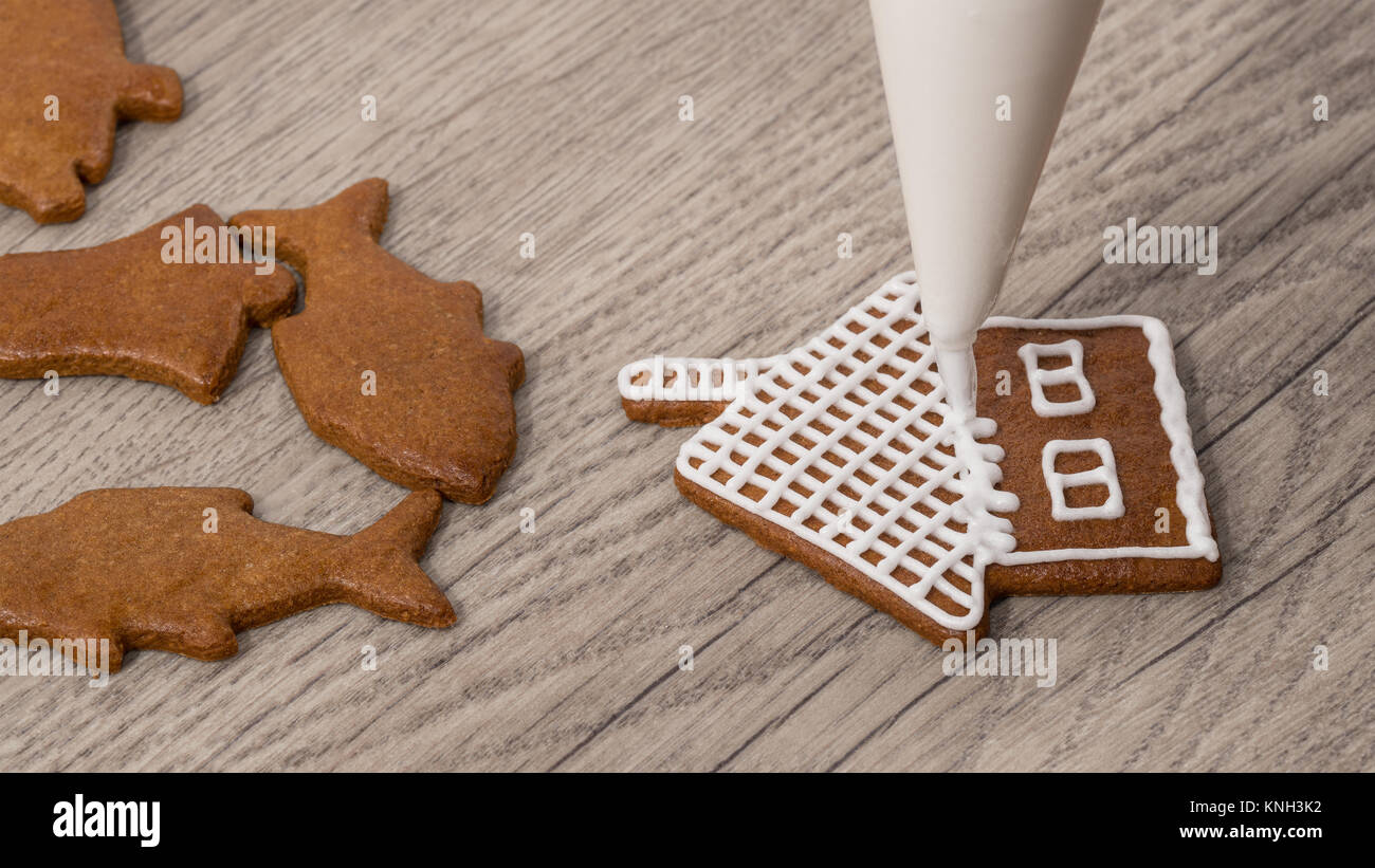 Close-up of decorating a christmas gingerbread in cottage shape. Painting of baked cookie using icing bag with sugar frosting on wood background. Stock Photo