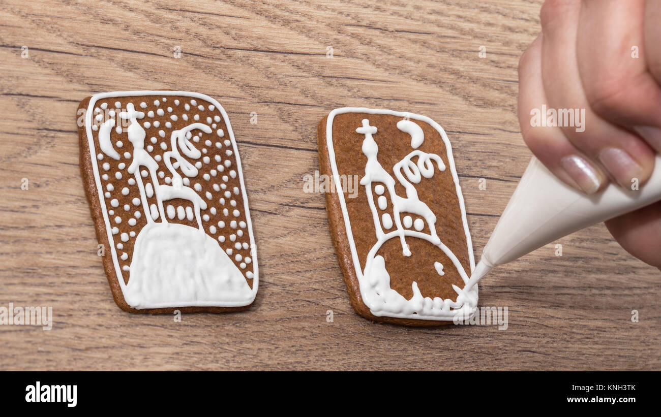 Close-up of female hand while decorating Christmas gingerbread on wood table. Painting the church on the biscuit using icing bag with sugar frosting. Stock Photo