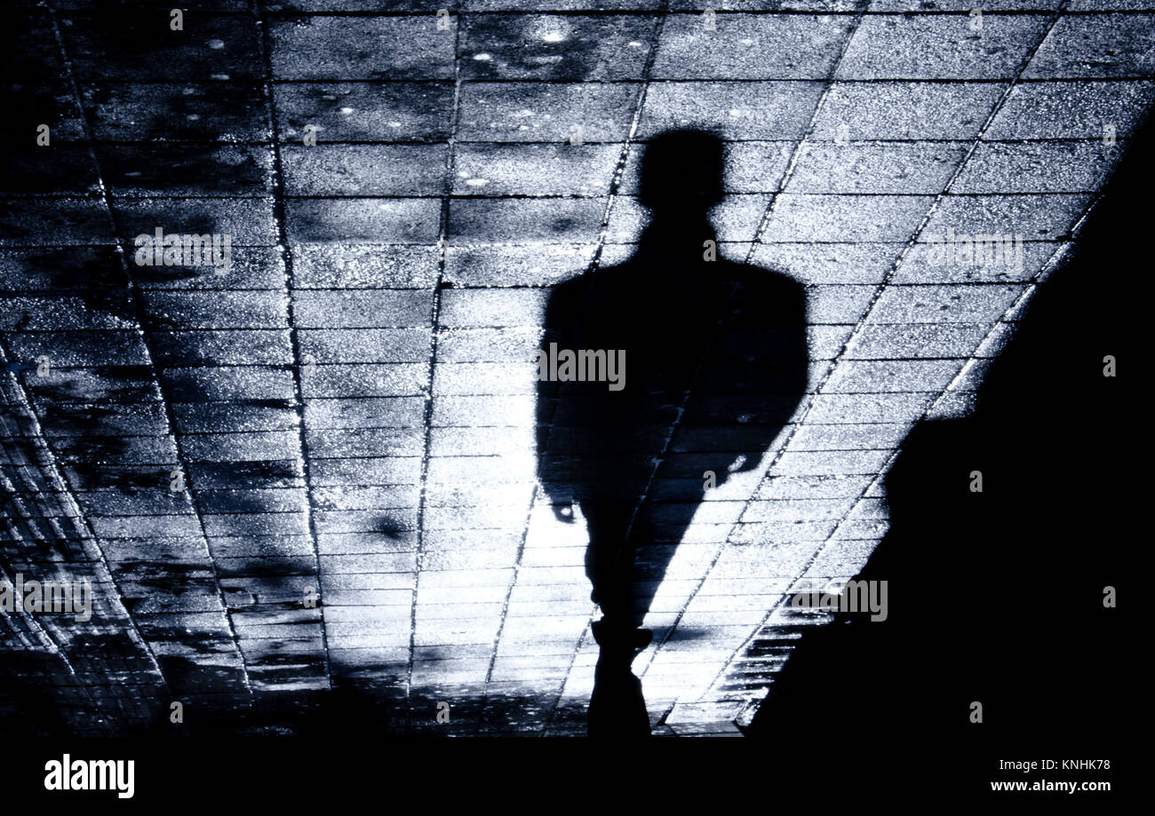 Blurry shadow and silhouette of a man standing in the night on wet city street sidewalk with water reflection Stock Photo