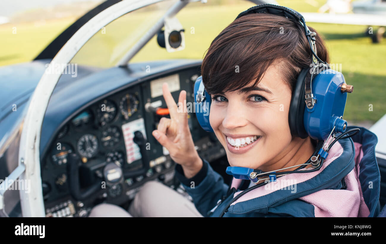 Smiling female pilot in the light aircraft cockpit, she is wearing aviator headset and making a V sign Stock Photo