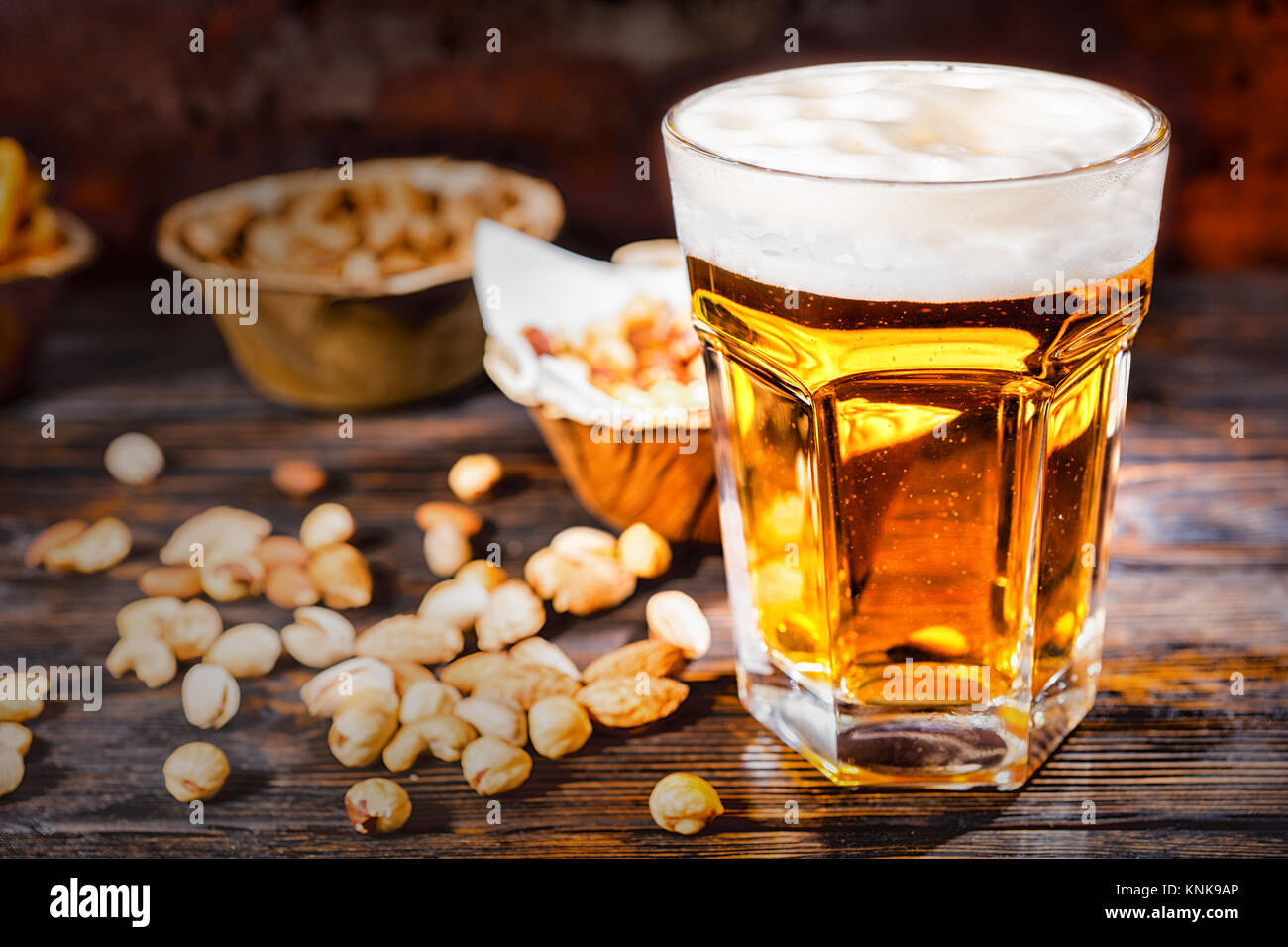 Glass with freshly poured light beer near plates with snacks and scattered nuts on dark wooden desk. Food and beverages concept Stock Photo