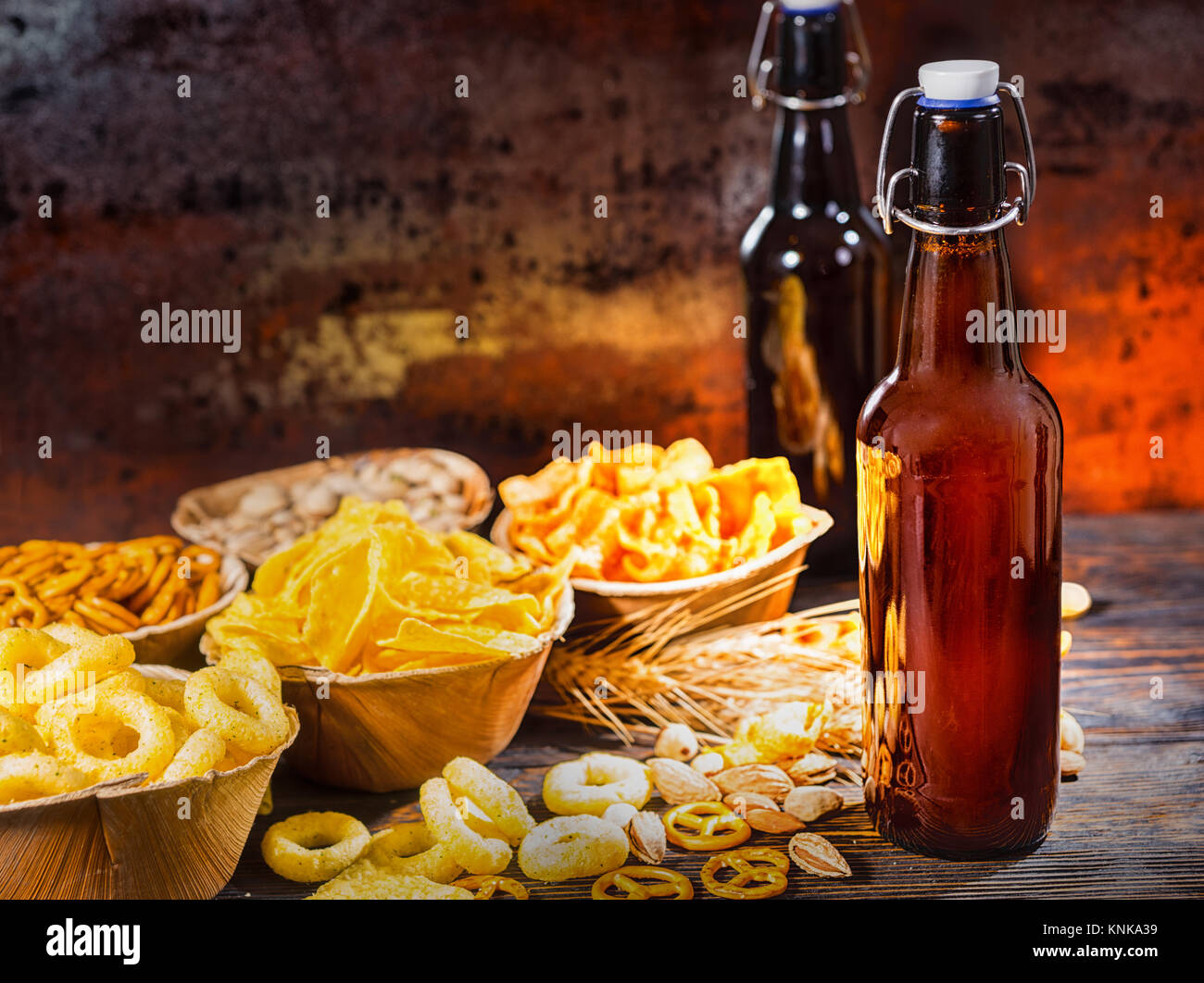 Plates with snacks near two bottles of beer, wheat, scattered nuts and pretzels on dark wooden desk. Food and beverages concept Stock Photo