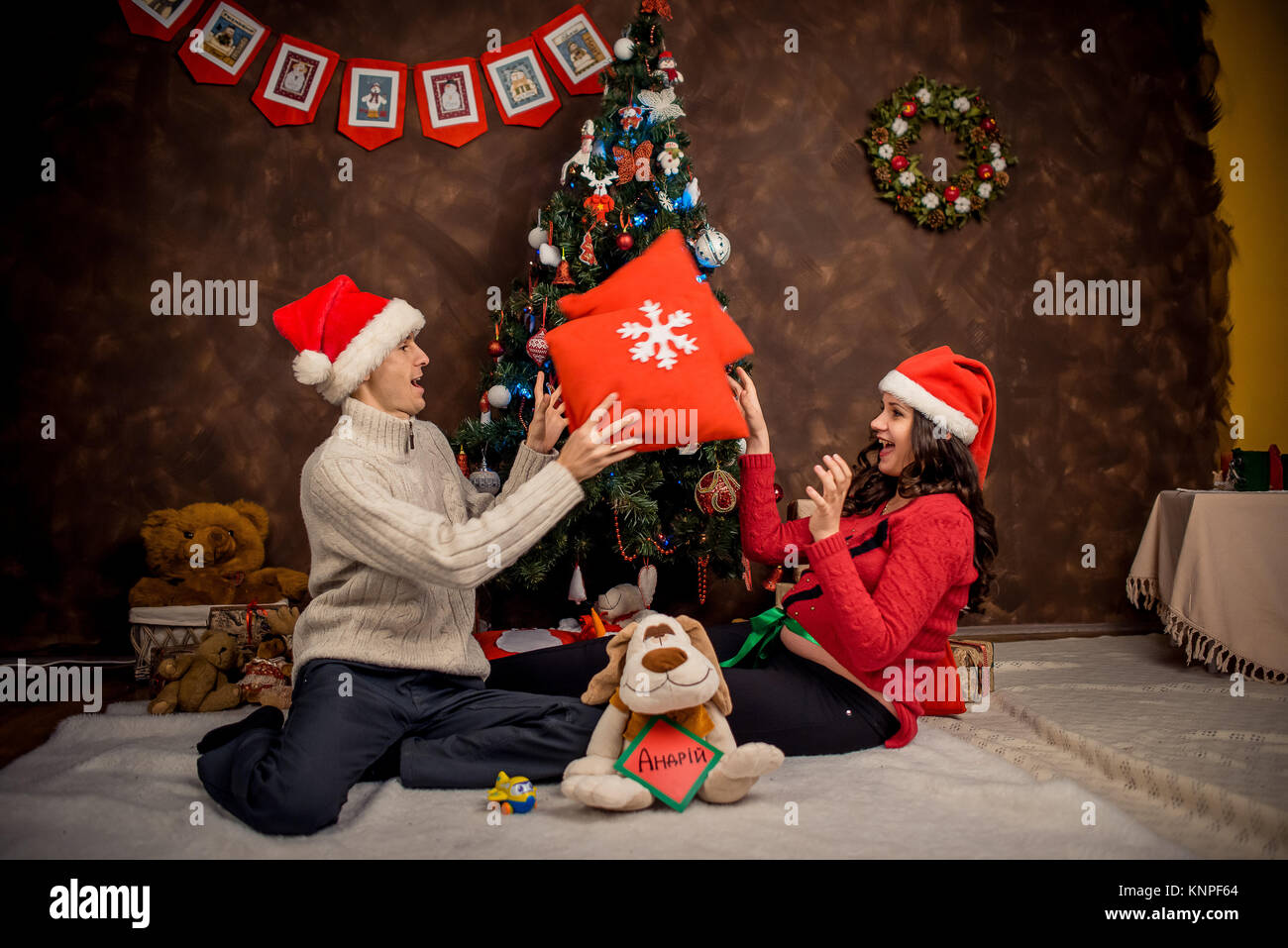 Smiling beautiful future parents are happily playing pillows while sitting on the floor near the decorated Christmas tree. Horizontal shot. Stock Photo
