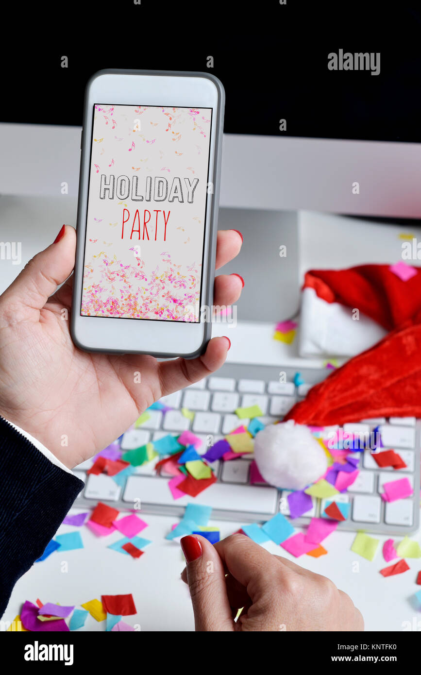 closeup of a young caucasian woman sitting at her office desk full of confetti looking to her smartphone, with the text holiday party in its screen Stock Photo