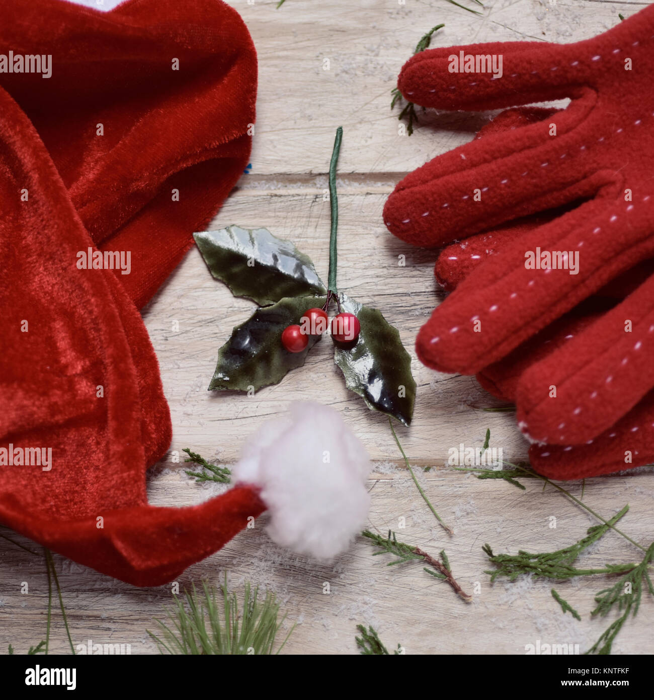 closeup of a santa hat, a pair of cozy red gloves, some twigs of pine tree and a twig of holly on a white rustic wooden surface Stock Photo