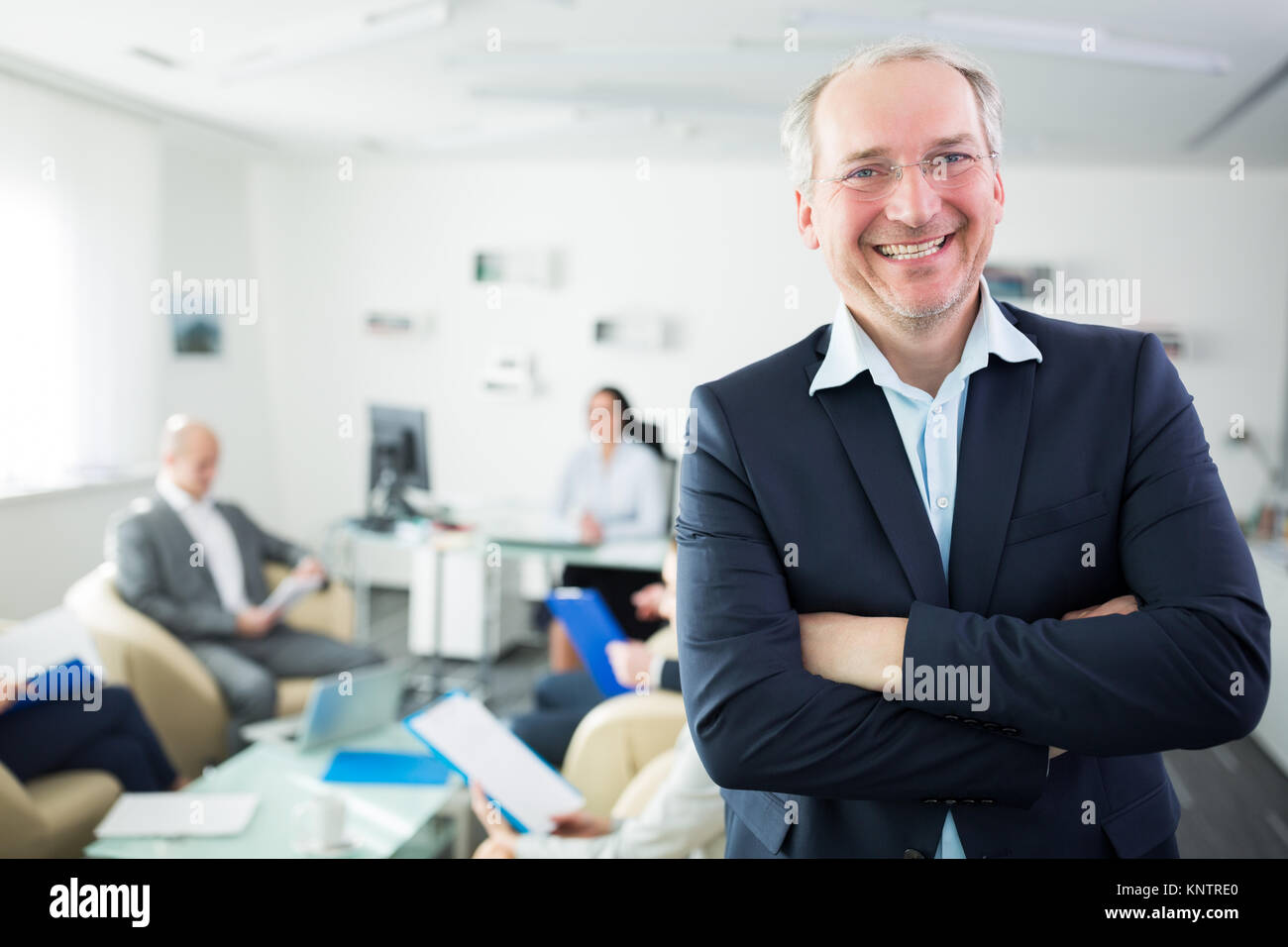 Portrait of smiling senior businessman standing arms crossed in office ...