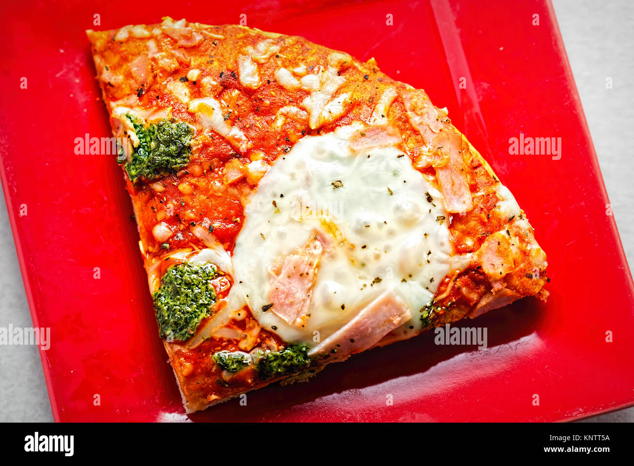 Slice of pizza with mozzarella, green pesto and ham on red plate Stock