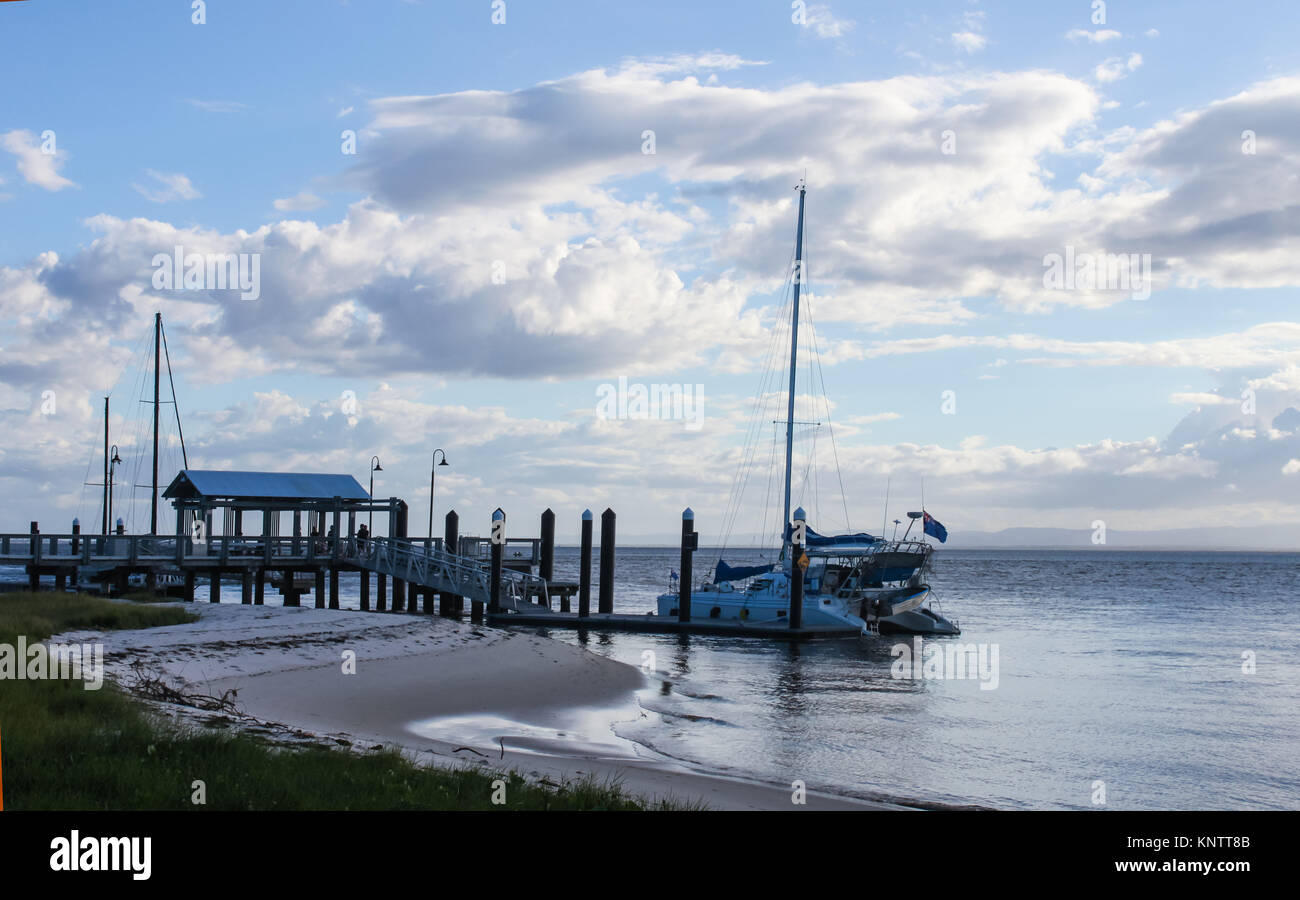 Dock in harbor at twilight with catamaran moored and other boats behind and people walking out of dock - mountains on the horizon and fluffy clouds in Stock Photo