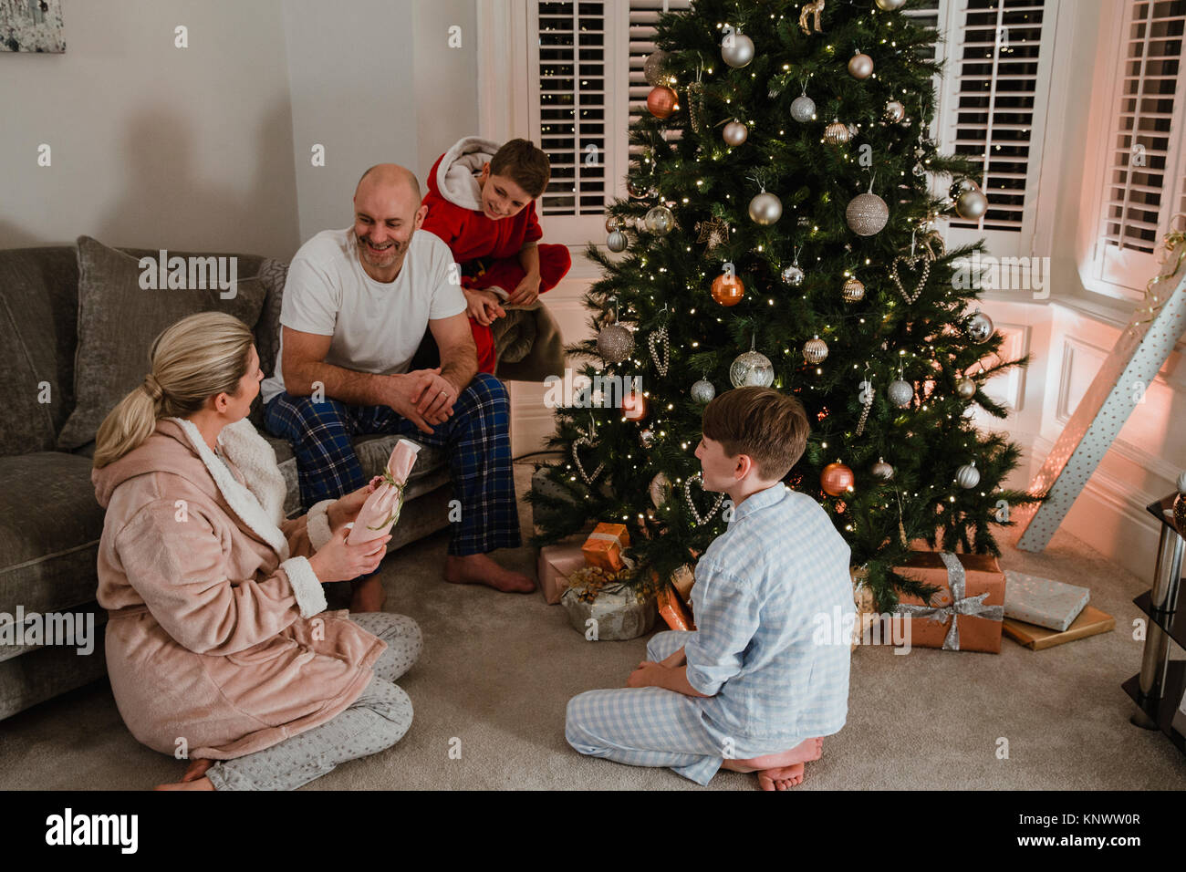Happy family are opening presents together on christmas morning. They are sitting by the christmas tree in the living room of their home, wearing pyja Stock Photo
